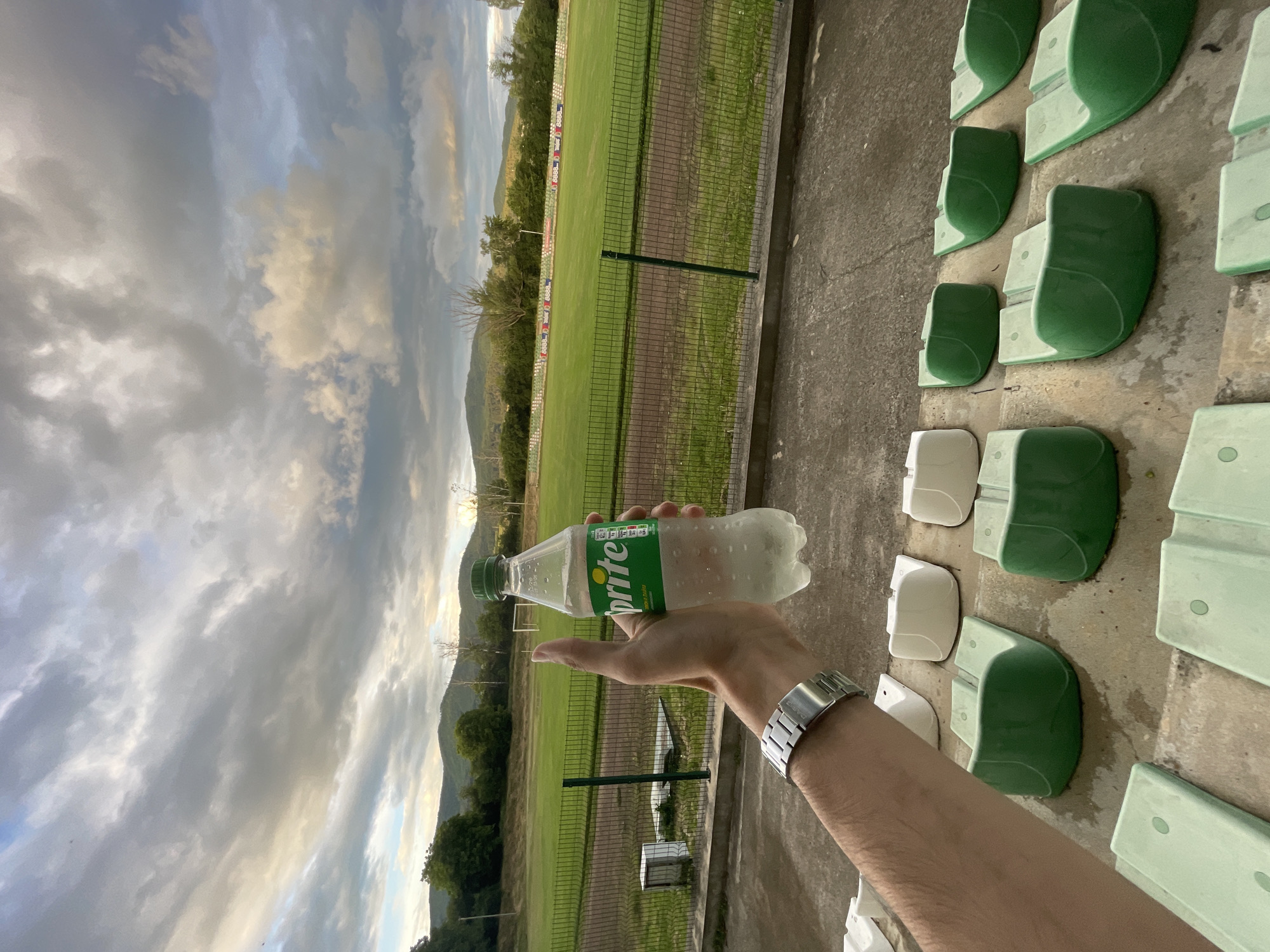 The image depicts an outdoor sports stadium or field in Ihtiman, Bulgaria, captured during either sunrise or sunset, indicated by the warm golden hues mixing with grey and blue clouds in the sky. In the foreground, a person's left arm and hand are prominently featured, holding a chilled plastic bottle of Sprite, visible with condensation and its distinctive green label. A metallic wristwatch with a link bracelet is worn on the person's wrist.
The scene includes a vibrant green grass field, bordered by a dirt track or running path. A green metal fence separates the field from the spectator area. In the distance, tree-covered hills or mountains form a natural backdrop. Along the far side of the field, various banners or advertisements are visible, with the number "8888" discernible on some sections. To the right, rows of stadium seating are present, consisting of concrete steps fitted with plastic seats in varying shades of green and some off-white. The field appears empty, suggesting a moment of quiet, perhaps before or after an event. The only clearly visible text is "Sprite" on the bottle label.