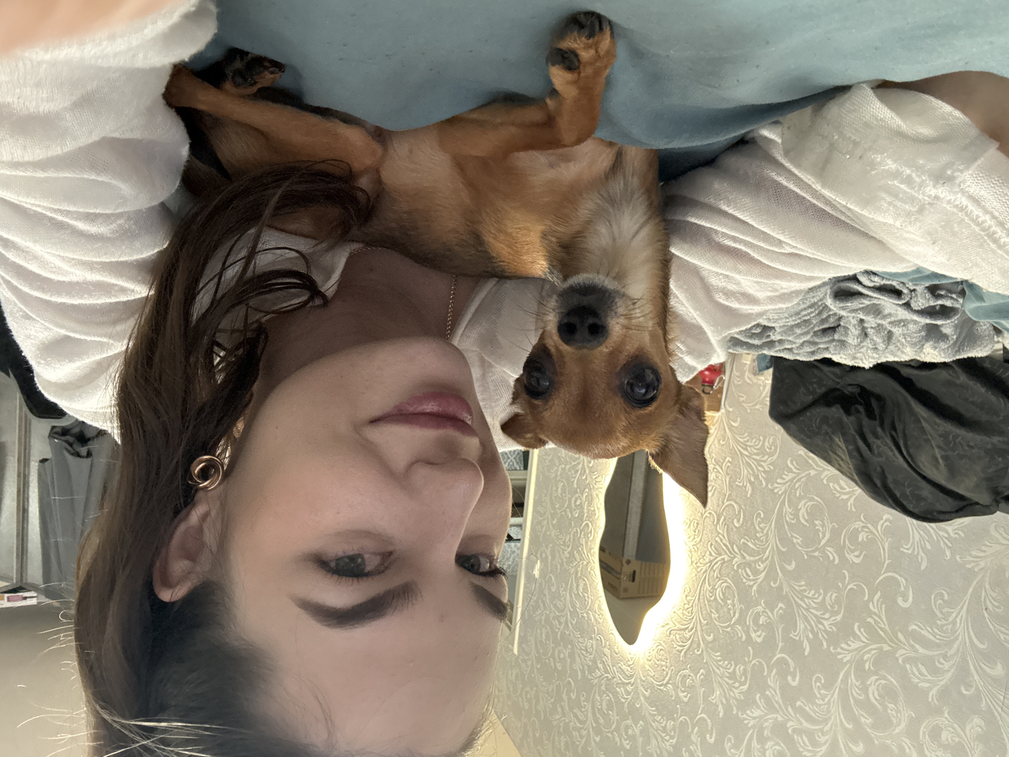This is a selfie of a young woman and a small brown dog, taken indoors. The woman is lying down, with her head angled towards the camera, and the dog is lying on top of her, also looking at the camera. The woman has long brown hair and is wearing a white top. The dog appears to be a chihuahua mix. The background shows a textured wallpaper and a lighted mirror. The overall mood seems casual and affectionate, with both subjects looking relaxed. The lighting suggests it is daytime, likely indoors with artificial lighting present. There is no visible text in the image.