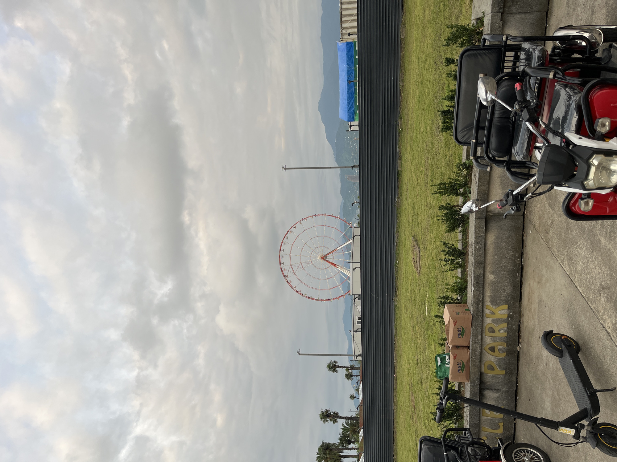 This image depicts an outdoor scene in Batumi, Georgia, likely near a park or recreational area. In the foreground, a black electric scooter is parked on a concrete surface. To its right, a red and black three-wheeled electric vehicle with two seats is also parked. Behind these vehicles is a low concrete wall with the words "CYCLE PARK" stenciled in yellow letters. On top of the wall are two cardboard boxes with the brand "muti" visible, and a green shopping bag.
The middle ground features a neatly manicured green lawn bordered by a dark, corrugated metal fence. Beyond the fence, a Ferris wheel with a red and white structure stands prominently against a cloudy sky. Palm trees are visible to the left of the Ferris wheel, and in the distance, mountains are shrouded in haze. A blue tarp covers some structures behind the fence, and shipping containers are also present. The sky is overcast with numerous clouds. There are no people visible in the image. The scene appears to be a public space, possibly a promenade or an amusement area.
