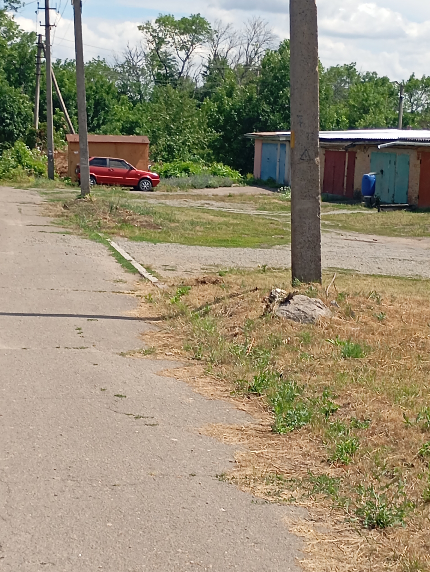 The image depicts a scene in Sozonivka, Ukraine, featuring a residential or industrial area with several garages and a red car parked near a brown shed. The foreground shows a paved road with patchy grass and dry vegetation along its edges. In the midground, a red sedan is parked to the left of a brown shed. To the right of the shed are multiple garages with colored doors, including blue, red, and teal. A large blue barrel is visible next to one of the garages. A concrete utility pole stands prominently in the midground, with another utility pole and some electrical wires visible in the background. The background is filled with dense green trees under a partly cloudy blue sky. The overall environment appears to be a quiet, possibly rural or suburban setting. There are no people visible in the image.