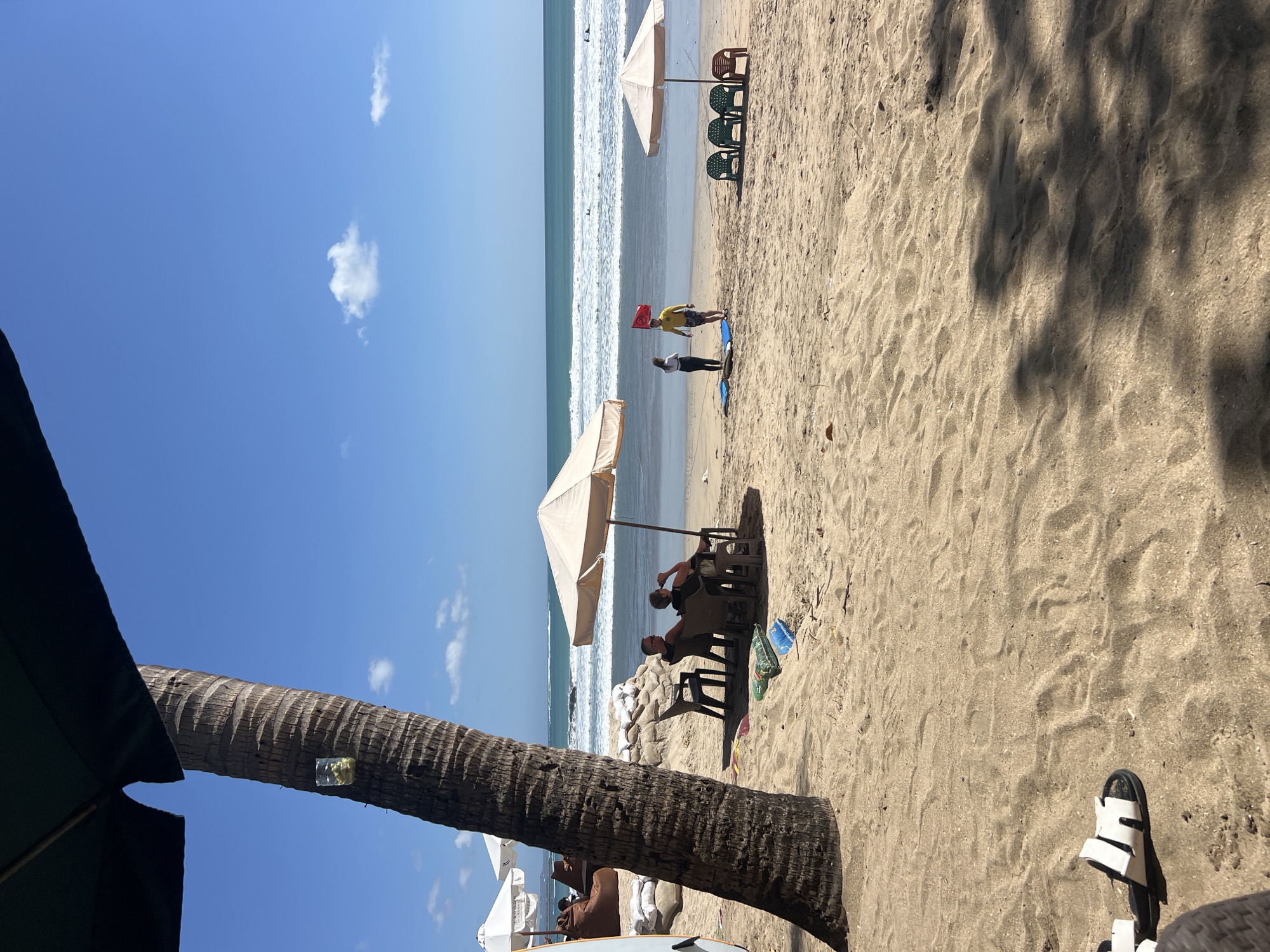 The image depicts a sunny day at Kuta Beach, Indonesia. A large palm tree dominates the foreground, with a glass containing what appears to be ice and possibly a drink tied to its trunk. Underneath the shade of an umbrella, two individuals are seated in chairs, facing the ocean. The sand is a light brown, with a few scattered items like bags and a blue plastic object.
Further down the beach, two people stand near a red flag, possibly a lifeguard or surf instructor and a student. Several empty green chairs are arranged nearby, along with a single brown chair. The ocean is calm with small waves, and a few surfers can be seen in the distance. The sky is a clear blue with a few scattered white clouds. The overall scene suggests a relaxed atmosphere at a popular tourist destination.
