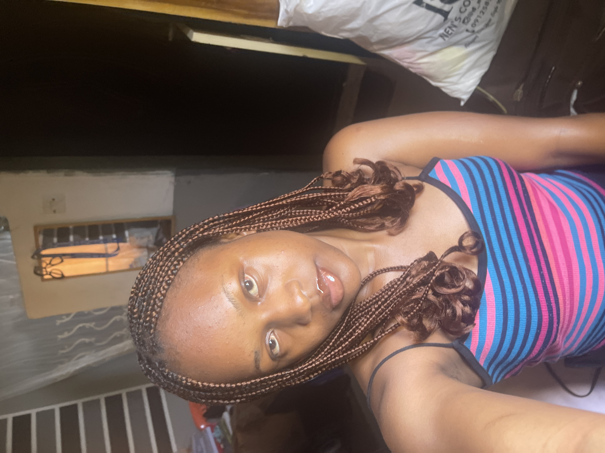 A young woman with braided brown hair is taking a selfie indoors. She is wearing a spaghetti strap tank top with horizontal stripes of pink, blue, and black. She has on lip gloss and a nose ring. The background is dimly lit and appears to be a room, with a dark doorway or closet visible to her right. A large white bag with some text and markings is also to her right. The wall behind her on the left is lined with dark horizontal slats. The overall impression is a casual, personal moment captured in what seems to be a residential setting. The specific location in Jos, Nigeria, cannot be determined from the visual information alone.
