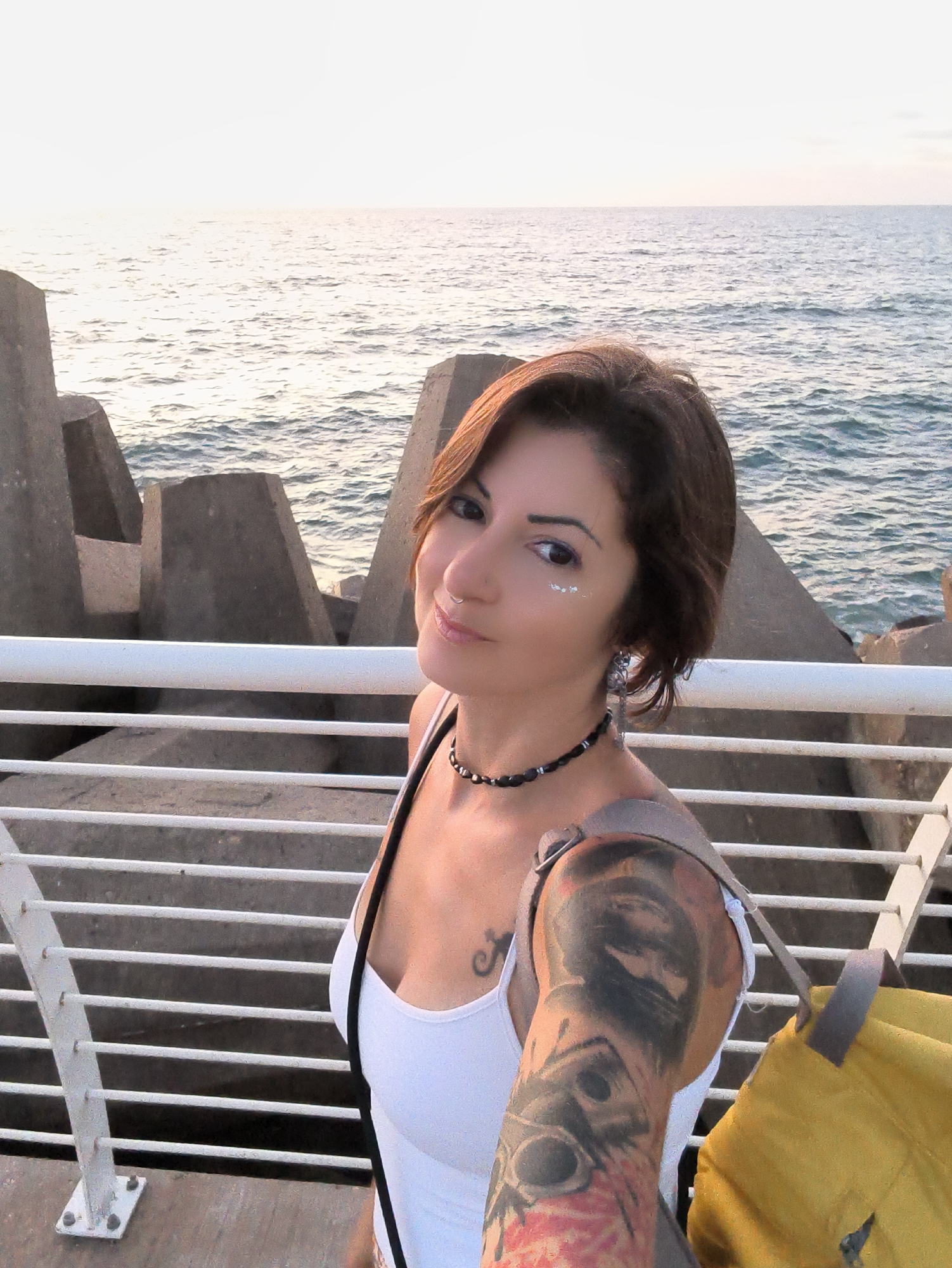 The image shows a woman with short brown hair and tattoos on her arm, standing on a walkway near the sea. She is wearing a white tank top and a black necklace. A yellow bag is slung over her shoulder. The background consists of the ocean, a breakwater made of concrete blocks, and a railing. The lighting suggests it is either sunrise or sunset. The specific location appears to be a promenade or pier in Ashkelon, Israel, given the architectural style of the breakwater and the general coastal environment.