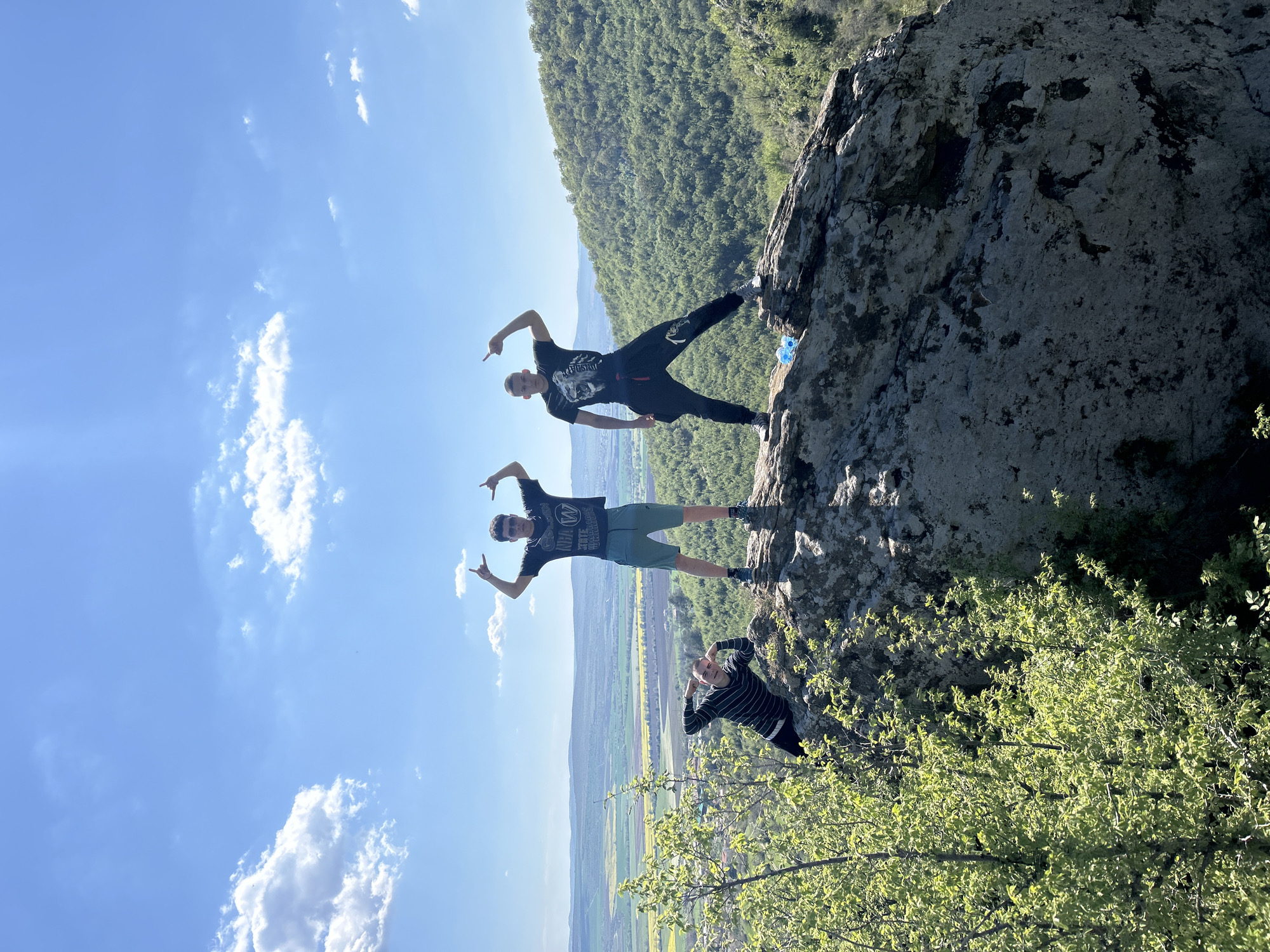 Three young men are standing on a rocky outcropping overlooking the landscape near Goritsa, Bulgaria. The man at the top is wearing a dark t-shirt, dark pants, and sneakers. He is pointing outwards with both hands. Below him, another man is wearing a dark t-shirt, light shorts, and sneakers and is also pointing outwards with both hands. At the bottom of the rock face, a third man is posing and wearing a horizontally striped dark and light colored shirt. They are near trees. In the background, there is a green landscape and blue sky with some clouds.
