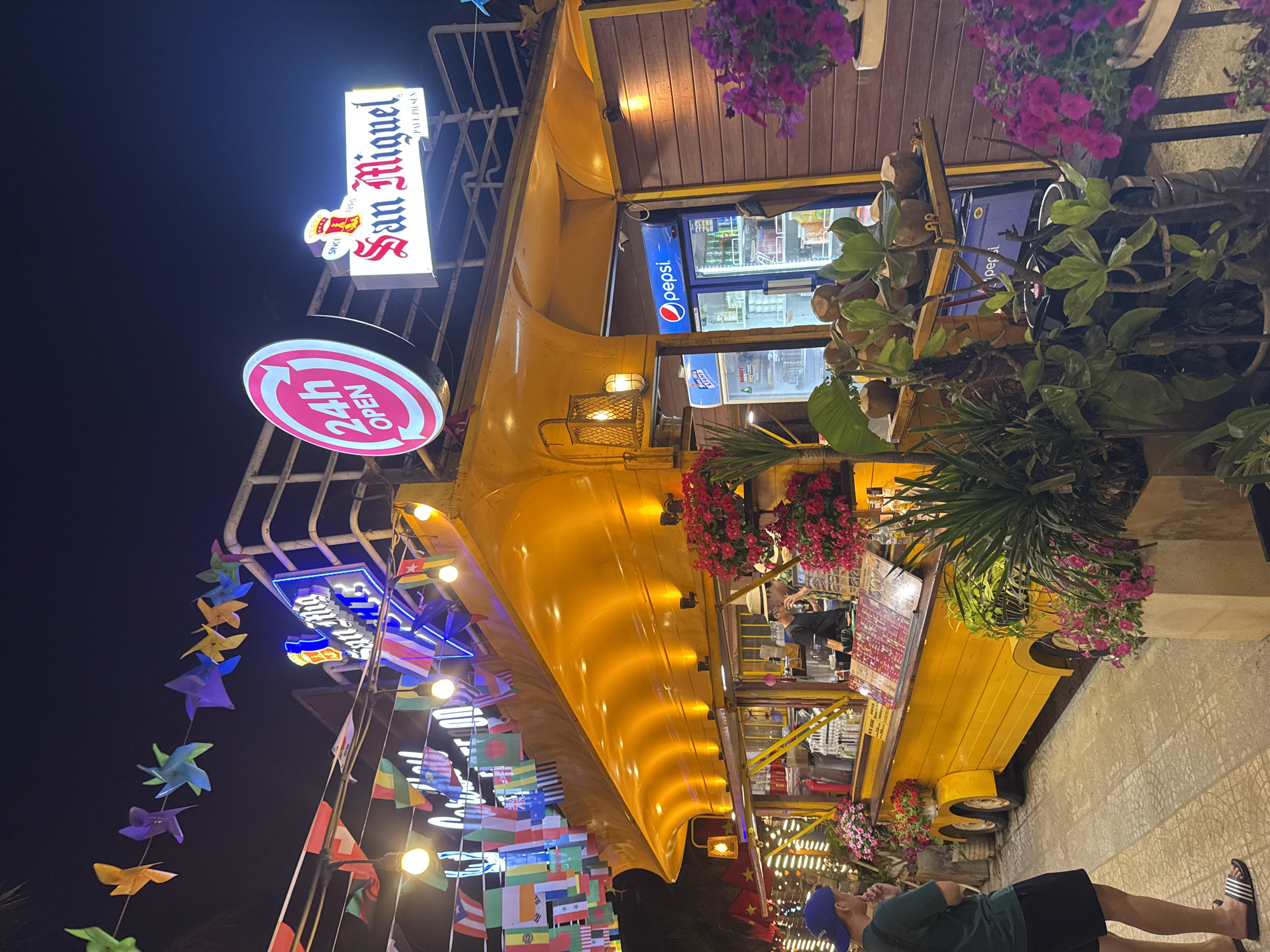 A brightly lit food establishment in Đà Nẵng, Vietnam, stands out against the night sky. The establishment is a converted yellow vehicle adorned with colorful pinwheels, international flags, flower planters, and various signs, including "San Miguel," "24h Open," and "Surf Five." A refrigerated Pepsi display is visible within the vehicle. A man in a dark green shirt, black shorts, and a blue baseball cap stands near the vehicle on a patterned tiled sidewalk.