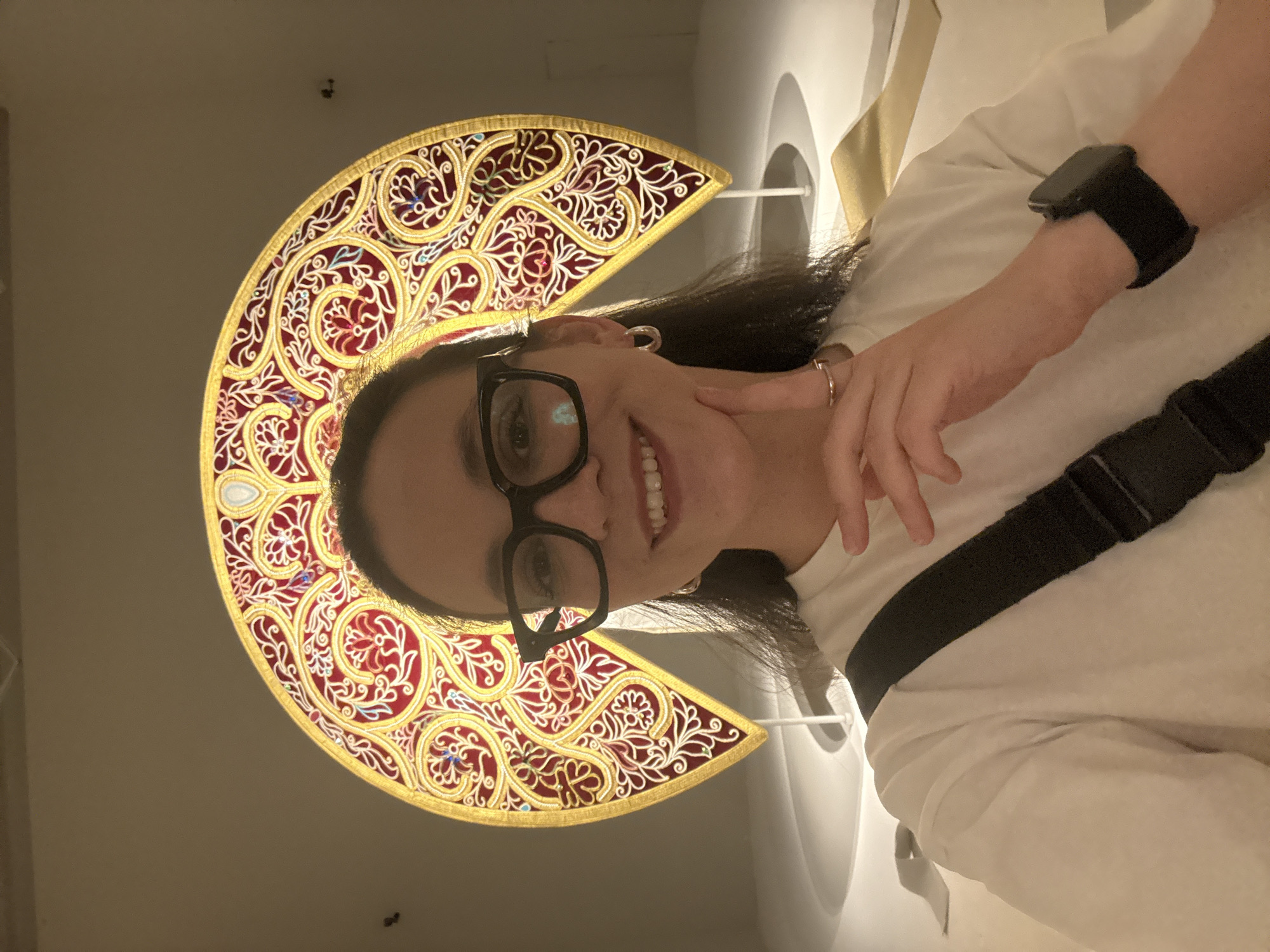 A woman with dark hair and glasses is standing in front of a decorative semi-circular artwork in Moscow, Russia. The artwork is red and gold with intricate designs. The woman is wearing a white shirt, a black watch, and a black strap across her body. She is smiling and holding her hand up to her face. The background is a blurred white or light-colored wall or ceiling.