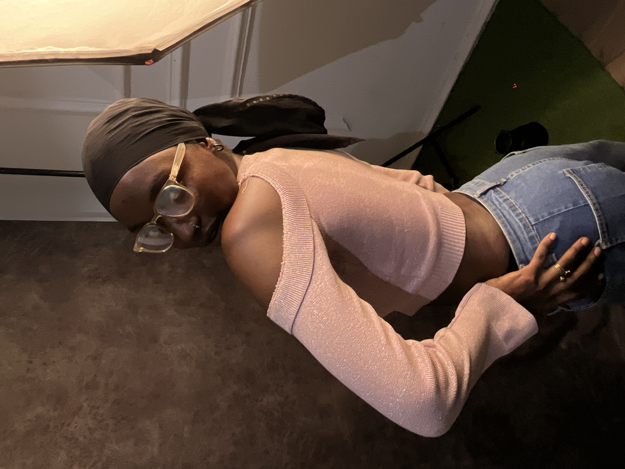 A person with dark skin and wearing a brown head wrap, gold-rimmed glasses, a pink, long-sleeved cropped top, and blue jeans is photographed from a low angle indoors. The individual is turned away from the camera, with their left hand visible on the jeans' waistband. Overhead, a softbox provides lighting. In the background, there's a glimpse of green, possibly foliage, and a black object. The location details are not discernable from the image alone.