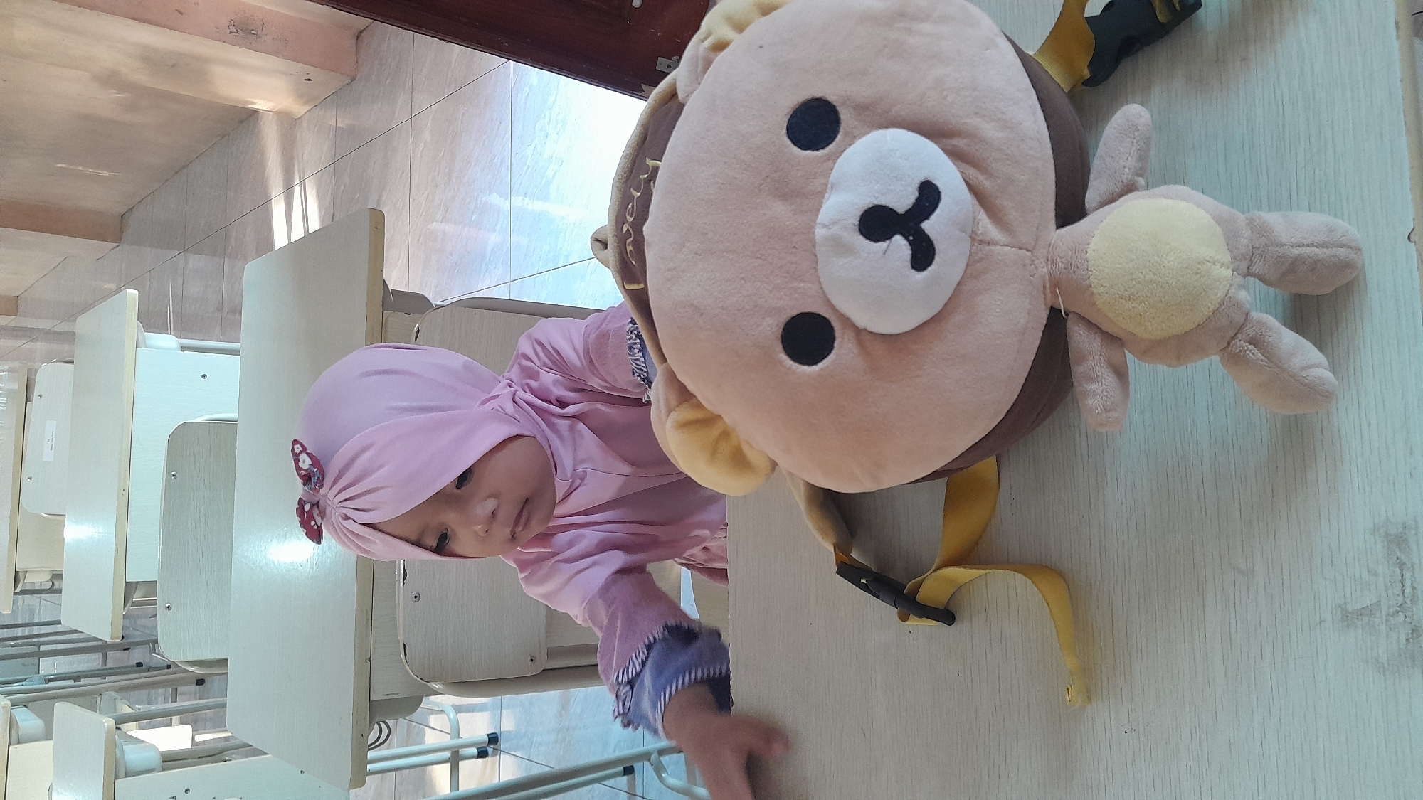 A young child wearing a pink hijab with a patterned bow sits at a desk in a classroom. The child is looking to their left. In front of the child on the desk is a large, plush teddy bear backpack. The classroom appears to be in a school, with rows of desks visible in the background. The lighting suggests it is daytime, and the floor is tiled. The image was likely taken in Tasikmalaya, Indonesia, given the context provided.