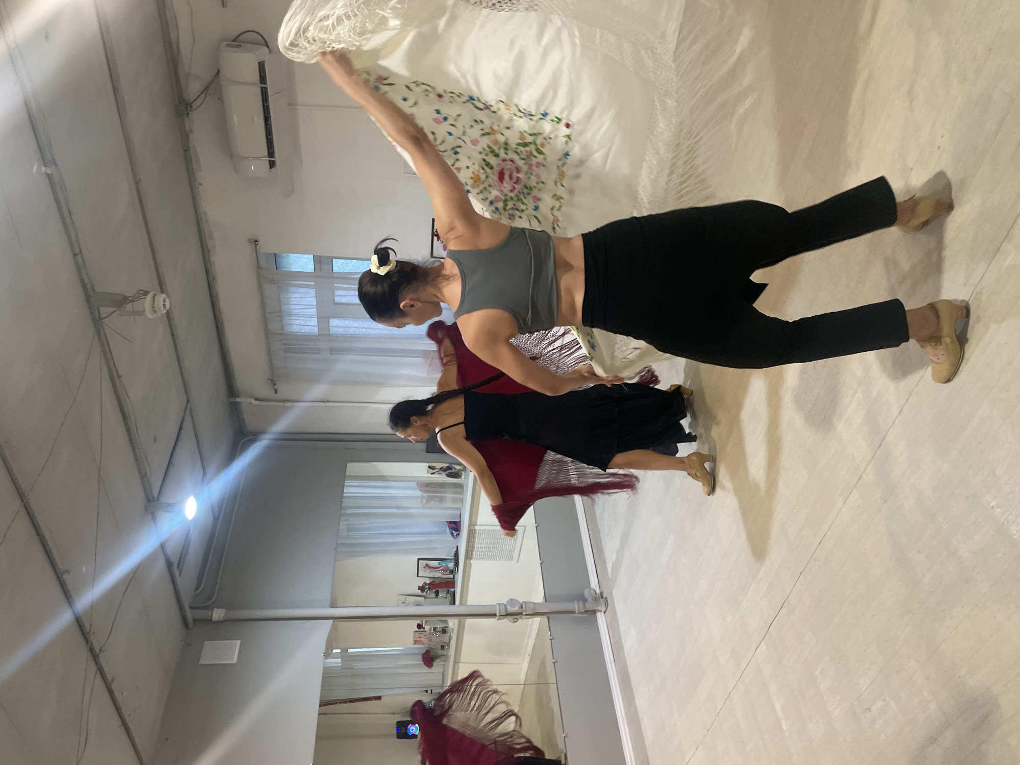The image depicts two women practicing flamenco dance in a studio. The woman in the foreground is wearing a grey sports bra, black loose-fitting pants, and beige heeled shoes. She is holding a white shawl with colorful floral embroidery, which she is swirling around her. The woman in the background is wearing a black flamenco dress and beige heeled shoes, and she is holding a dark red fringed shawl. The studio has a polished wooden floor, large mirrors on one wall, and white curtains on another. The ceiling is industrial, with exposed pipes and lighting fixtures. The overall atmosphere is one of active movement and artistic expression. The image does not contain information indicating the location is Sochi, Russia.