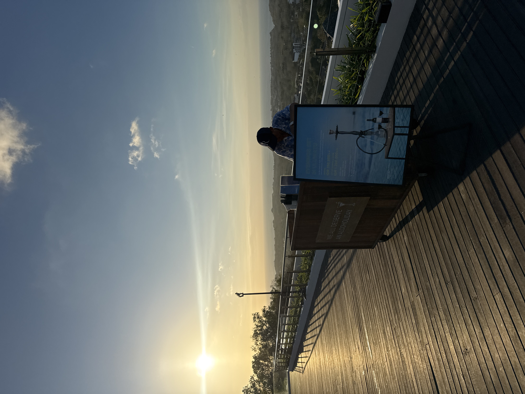 A man wearing a baseball cap and a patterned short-sleeved shirt leans over a wooden bar cart. The cart has wheels and a sign attached to its front displaying an image of a hookah, two wine glasses, and a bottle of wine, along with some text. To the left of the bar cart is a wooden deck illuminated by the setting sun, casting long shadows. In the background, a metal railing lines a walkway, beyond which is a landscape of trees, buildings, and the ocean. The sky is a clear blue with a few wispy clouds, and the sun is low on the horizon, creating a bright glow. This scene appears to be at a vantage point overlooking the coast of Kuta Selatan, Indonesia.