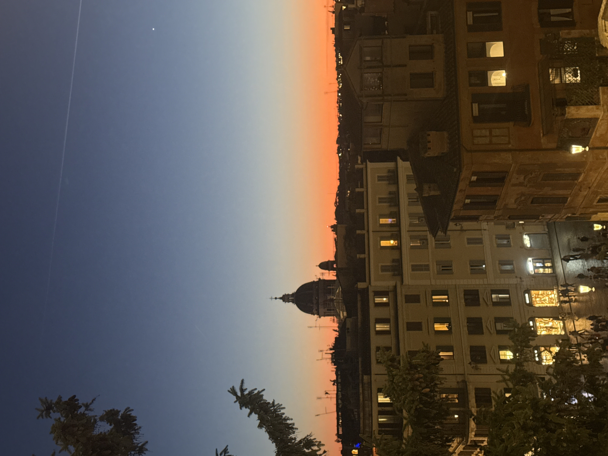 The image shows an urban landscape in Rome, Italy, at twilight. The sky transitions from blue to orange near the horizon, with a white dot, probably a star, visible. A straight line across the sky could be an airplane contrail. In the center of the image, the dome of a prominent building, likely St. Peter's Basilica in Vatican City, stands out against the sky.
The right side of the image depicts buildings, with windows lit up, suggesting it's evening. There are people on the street below, which appears to be slightly wet, possibly from rain. The overall impression is that of a cityscape at dusk, with a mixture of natural light and artificial illumination. There are trees in the foreground.