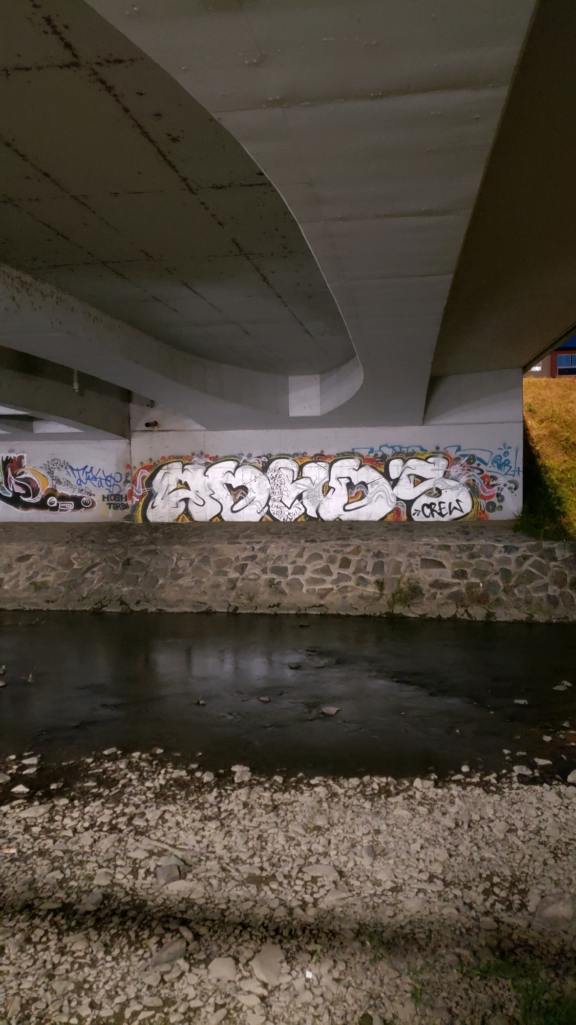 The image depicts graffiti on a concrete wall beneath a bridge, with a river flowing in the foreground. The graffiti style is bold and colorful, featuring a large, white bubble-lettered tag and smaller, more intricate designs. The wall itself is somewhat worn and stained, suggesting it is a public space. The river is dark and appears shallow, with a rocky bed. The overall atmosphere is one of urban decay and artistic expression. The location is confirmed to be under the bridge on the road Klatovská in Prešov, Slovakia.