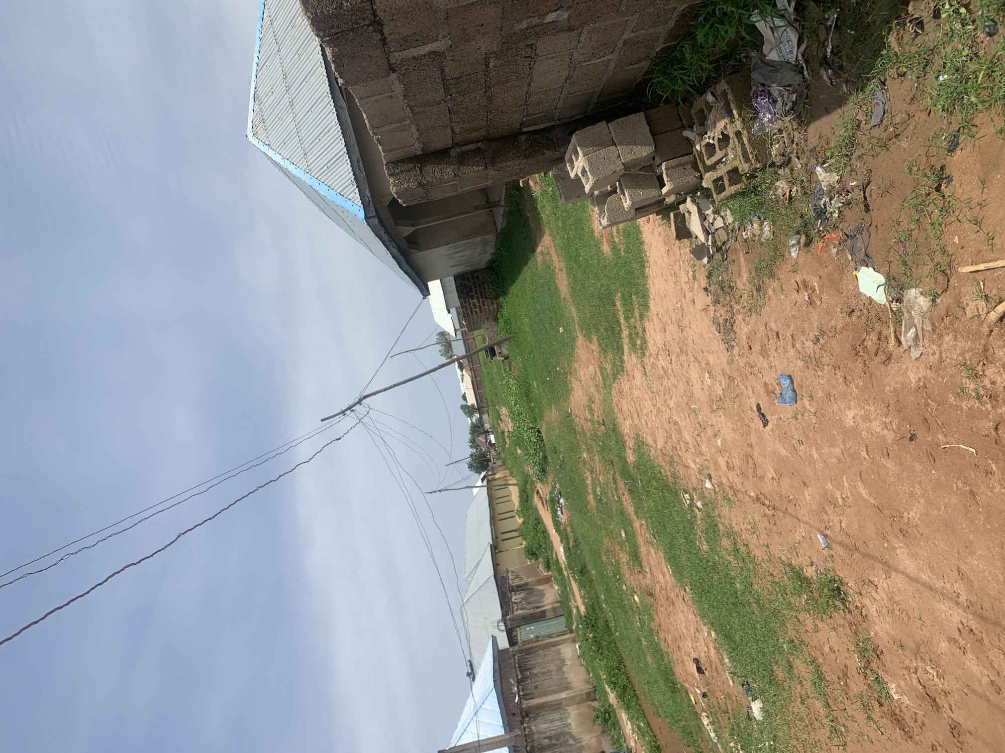 The image depicts a scene in the city of Jauro Yinu, Nigeria. In the foreground, a dirt path, littered with plastic bottles and other debris, winds its way through sparse green grass. To the right, a wall constructed of concrete blocks stands tall, with a stack of loose blocks and more refuse piled at its base. Beyond the wall, a building with a corrugated metal roof, trimmed with blue, is visible. Further into the background, a row of houses with similar corrugated roofs lines a street, with overhead power lines crisscrossing the sky. The sky is overcast with light clouds. The overall impression is one of a residential area undergoing development or with ongoing construction.
