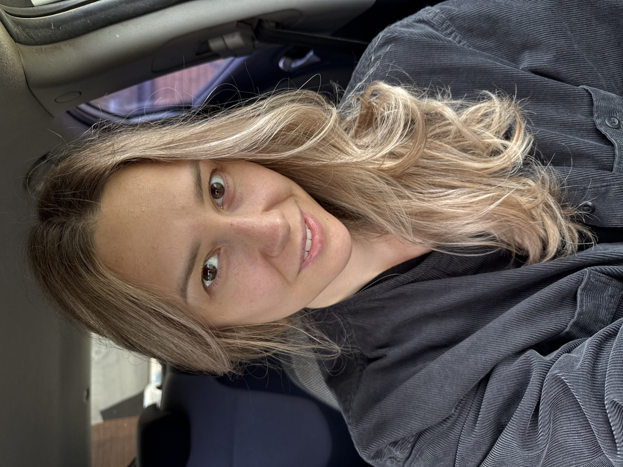 The image features a close-up portrait of a woman, seemingly taken from a slightly low angle, seated within what appears to be a car. She has long, wavy blonde hair with visible darker roots, and warm brown eyes that look directly at the camera. A gentle, friendly smile plays on her lips, revealing a glimpse of her teeth. She is dressed in a dark, ribbed fabric shirt or jacket, possibly corduroy, worn over a darker underlying top.
The setting is primarily the interior of a vehicle, with parts of the car's light-colored headliner and a dark window frame visible. Through the car window on the left side of the frame, a blurred outdoor background can be seen, hinting at a building or wall under natural daylight. The woman's expression conveys a sense of ease and approachability. There is no visible text in the image, and the lighting suggests it is daytime.