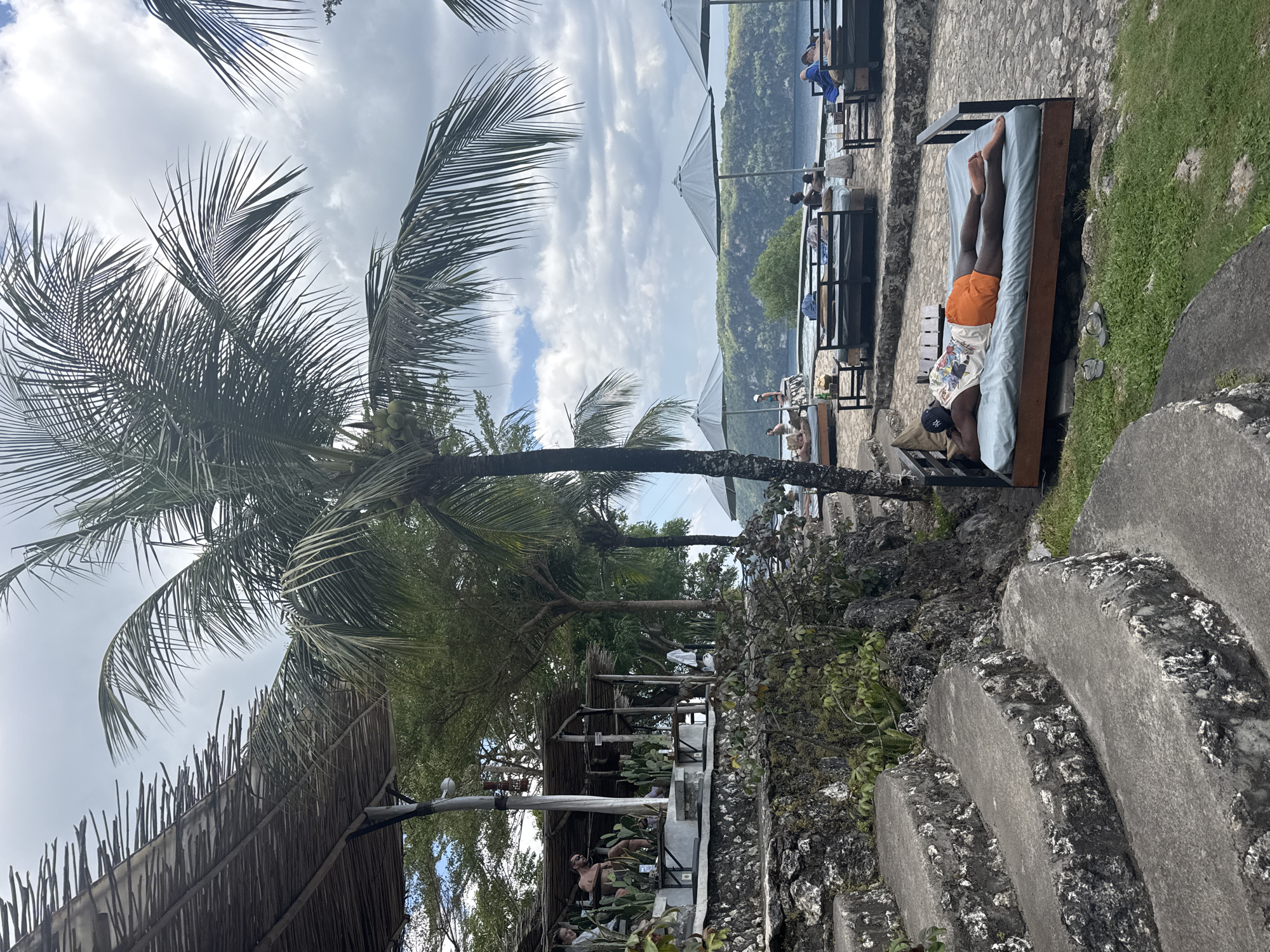 This image captures an outdoor leisure area, identified as being in Nusa Penida, Indonesia. The scene is characterized by terraced architecture, lush tropical vegetation, and a view of a body of water with a distant landmass.
In the immediate foreground, a set of roughly hewn, dark grey concrete steps with white, textured edges curves upwards from the bottom left. To the right of these steps, a dark-skinned individual lies face down on a light blue mattress, which rests on a dark wooden lounge bed. This person wears orange shorts, a light-colored graphic tank top, and a dark baseball cap. A pair of grey flip-flops is positioned on the small patch of green grass beside the bed. The ground surrounding the lounge bed is paved with irregular dark grey stones.
In the midground, a prominent palm tree extends upwards from the lower center, displaying numerous green fronds and a cluster of green coconuts. To the upper left, an open-air structure with a thatched roof and wooden supports is visible. Inside this structure, several individuals are seated at tables amidst large cactus plants. One man with a beard is clearly visible, looking right.
Beyond the foreground lounge bed and the main palm tree, a paved area hosts additional dark grey lounge beds with light blue mattresses, shaded by several large, circular grey umbrellas. Multiple individuals are seen lounging on these beds or sitting near the edge of a retaining wall overlooking a calm, blue-green body of water. One person in a blue swimsuit sits on the wall's edge.
The background features a wide expanse of partly cloudy sky with bright white cumulus clouds against a lighter blue. Across the water, a significant landmass with dense green vegetation and an undulating ridge line forms the distant horizon.
