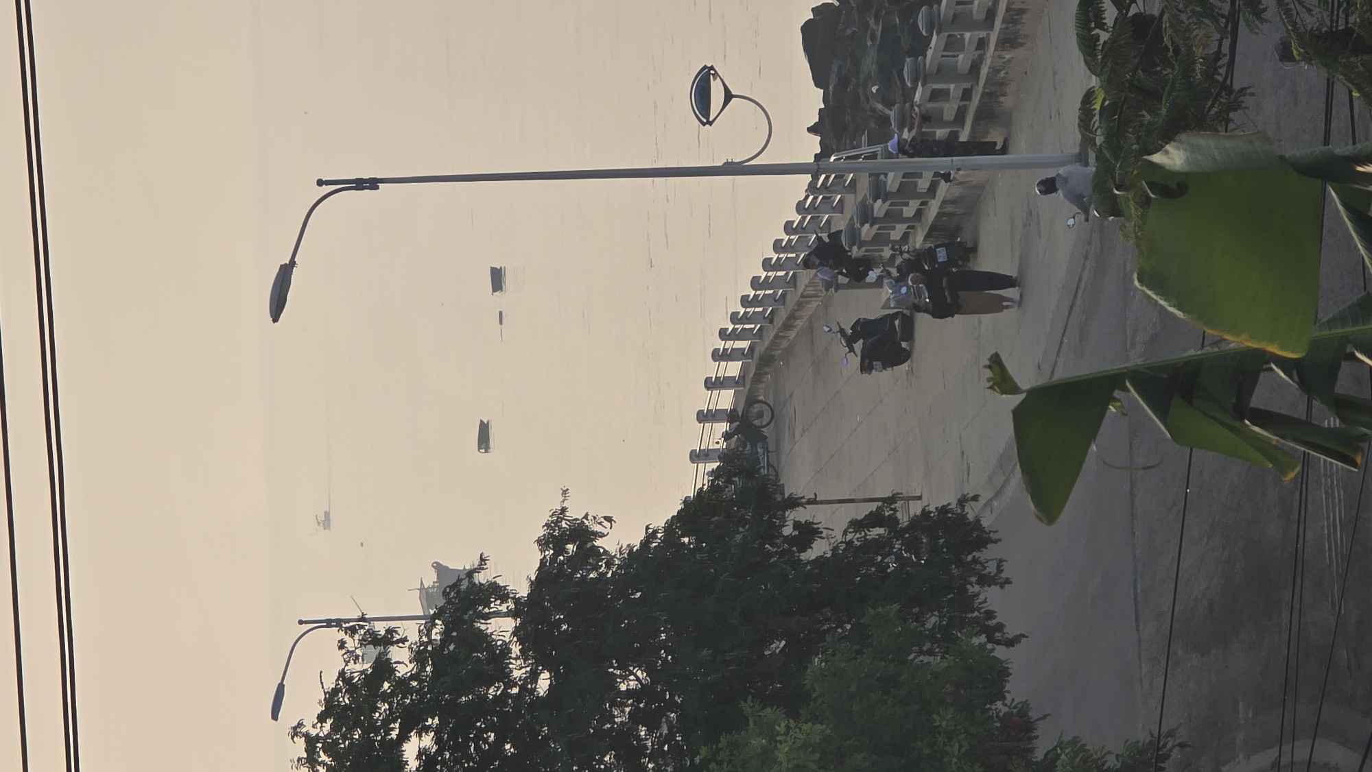 The image depicts a waterfront scene in Bac Natural Heritage Area Trang, Vietnam, taken from an elevated perspective. The sky is overcast. A concrete embankment runs along the shoreline, with several motorcycles parked along the road next to it. Multiple boats are visible on the water. Streetlights are present along the roadside, and trees and other vegetation are on the right side of the image. Power lines run across the left side. Some individuals are present on the embankment.