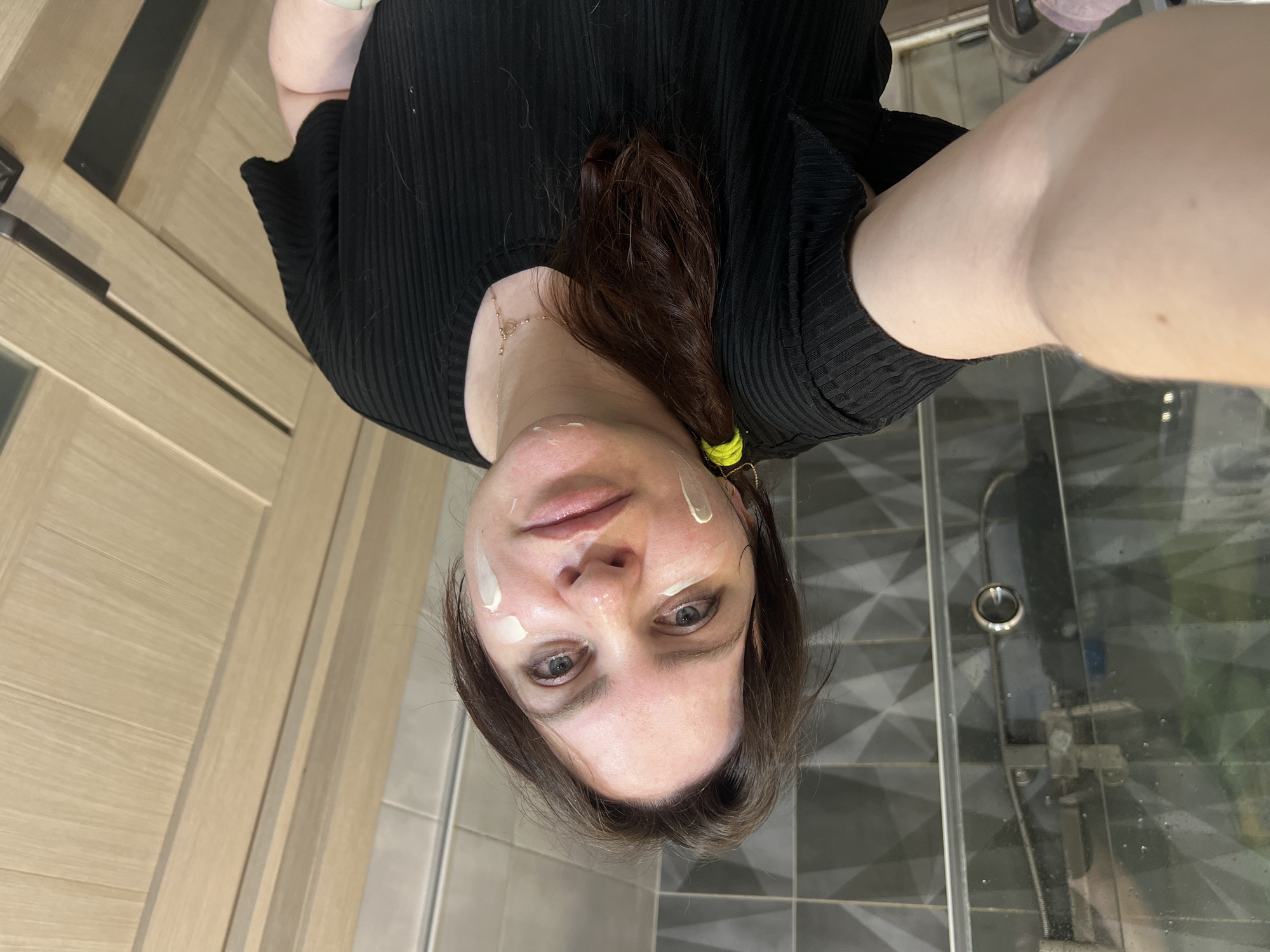 A woman is taking a selfie in what appears to be a bathroom. She has fair skin, brown hair pulled back into a ponytail with a yellow hair tie, and some white cream or lotion applied to her face. She is wearing a black ribbed top and a gold necklace. The background features a shower stall with grey tiles, a clear glass door, and a showerhead. A wooden door is visible to the left of the frame. It is impossible to ascertain the specific location in Noviy, Russia, based on the image.