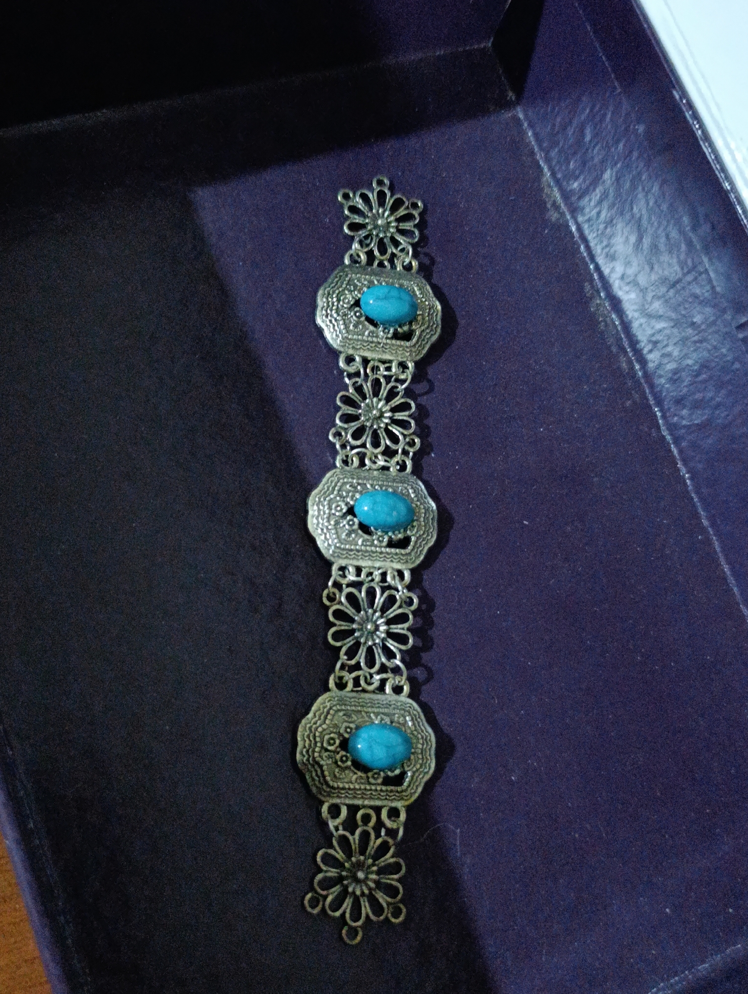 A close-up, high-angle shot shows an ornate, silver-toned bracelet with three oval, turquoise-colored stones. The bracelet is laid vertically within a dark purple, open box. The box has a textured surface and appears to be made of cardboard or a similar material. The background is dark and out of focus. The image does not contain any information about the city of Manghit, Uzbekistan.