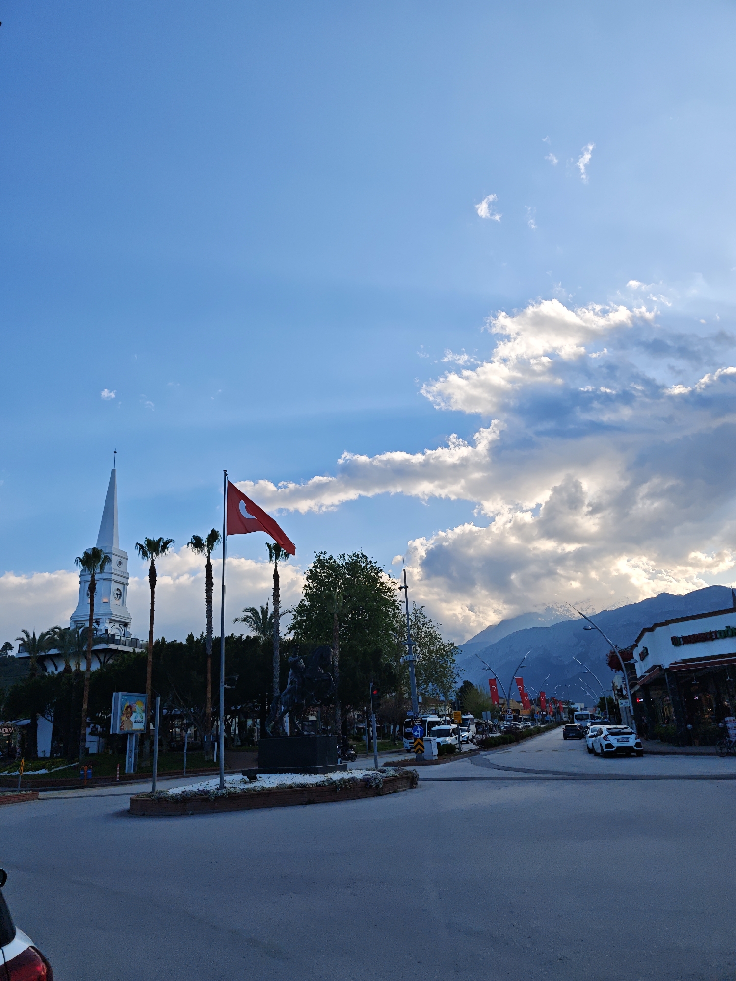The image shows a street scene in Kemer, Turkey. In the foreground, a street intersection is visible with cars and other vehicles driving on it. A roundabout is also present, featuring a statue and a Turkish flag. In the background, there is a building with a white spire, possibly a mosque or landmark structure. Palm trees and other vegetation are interspersed throughout the scene. The sky is partly cloudy with blue patches and sunlight shining through. Mountains are visible in the distance.