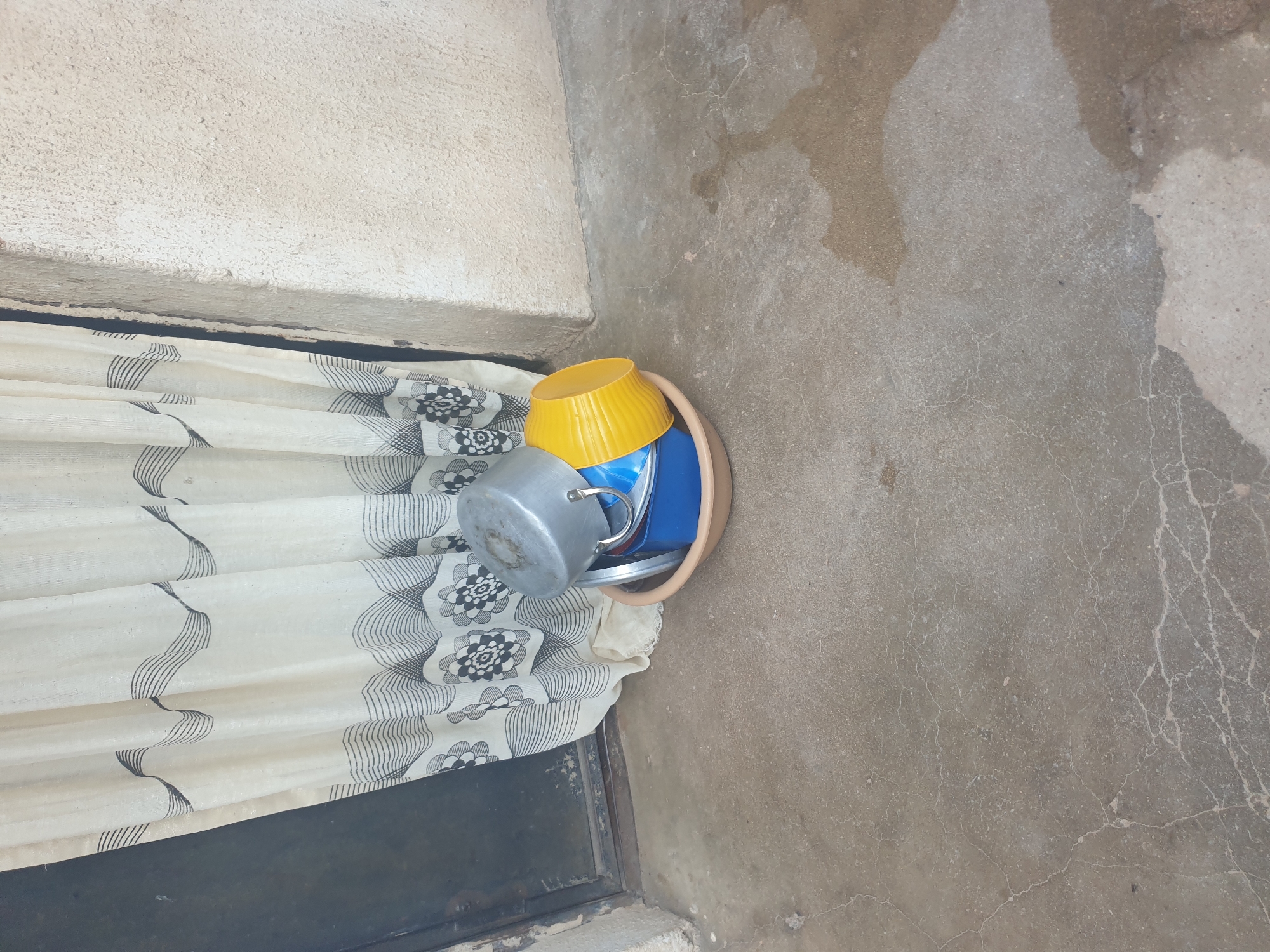 In the city of Unknown, Unknown, on a concrete surface, a stack of kitchenware is placed next to a wall and a curtain. The kitchenware includes a beige basin, a blue container, a yellow bowl, and a metallic pot. The basin, which appears to be made of plastic, is round and shallow. Inside it, a blue rectangular container is visible, followed by a yellow bowl, and finally a metallic pot. The wall behind the kitchenware is textured and painted in a light color. A cream-colored curtain with a black wavy pattern and floral motifs hangs on the left side of the scene, partially covering a dark doorway. The concrete ground is slightly wet and appears to have cracks.
