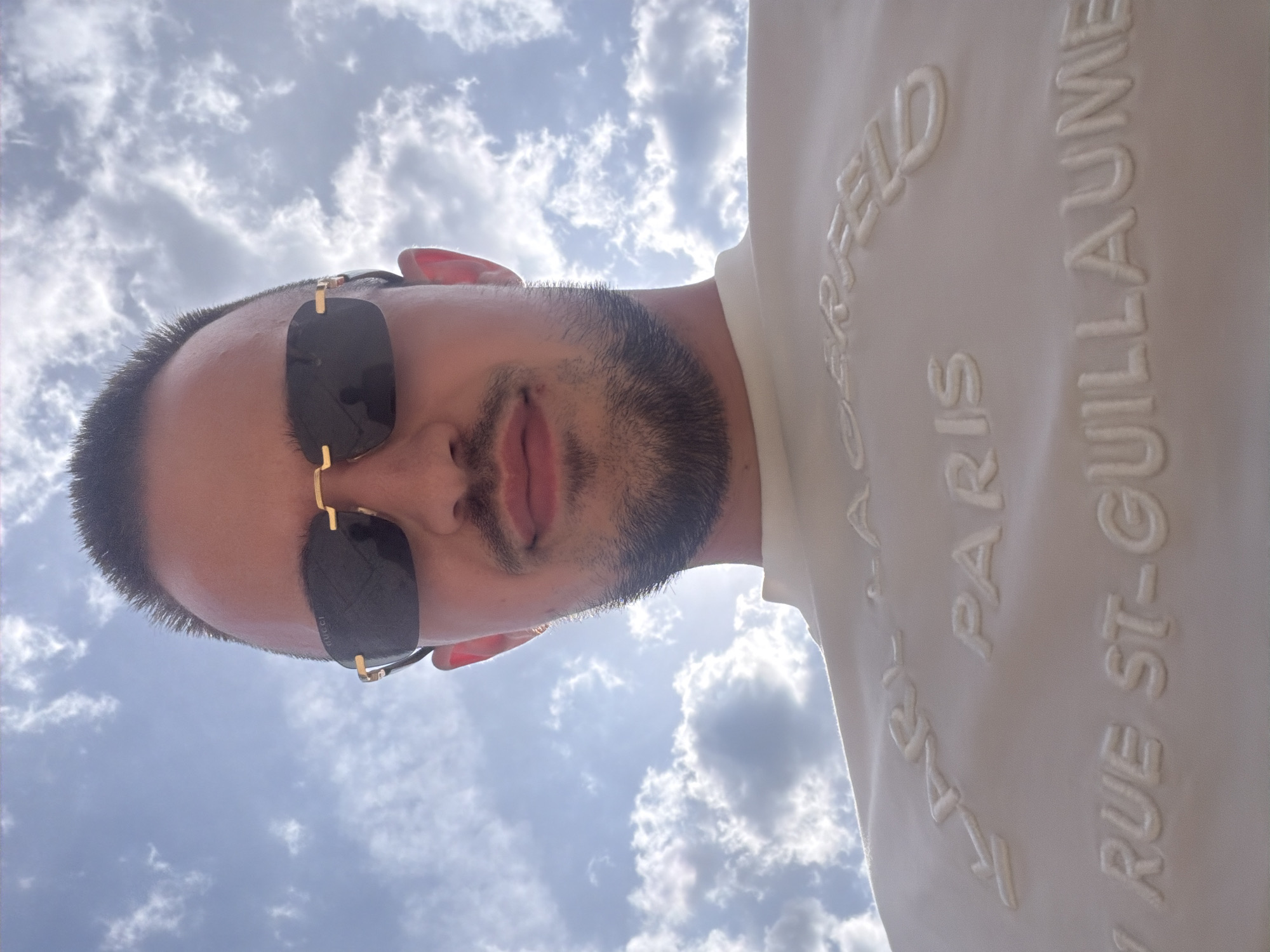 A low-angle, medium shot shows a young man with short, dark hair, a short beard, and a light complexion. He is wearing dark, rimless sunglasses with gold accents and a white t-shirt with raised lettering. The text on the t-shirt appears to be "KARL LAGERFELD PARIS RUE ST-GUILLAUME". He is looking upwards towards a cloudy sky. The image is shot outdoors. The location is not specified in the image, and therefore cannot be determined to be Sofia, Bulgaria.