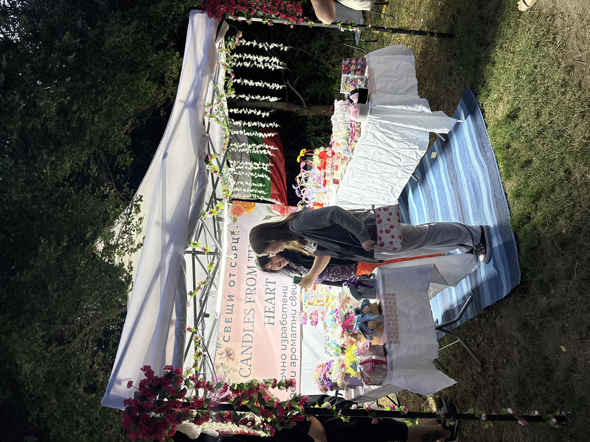 At dusk, outdoors, a market stall displays handmade candles and gift items. The stall is covered by a white canopy and decorated with floral garlands. Two women are standing at the stall, with one examining a product. Various gift boxes, baskets filled with decorative items, and small pastries are arranged on a white tablecloth. A Bulgarian flag hangs in the background. The ground is covered with grass and a blue striped mat.
