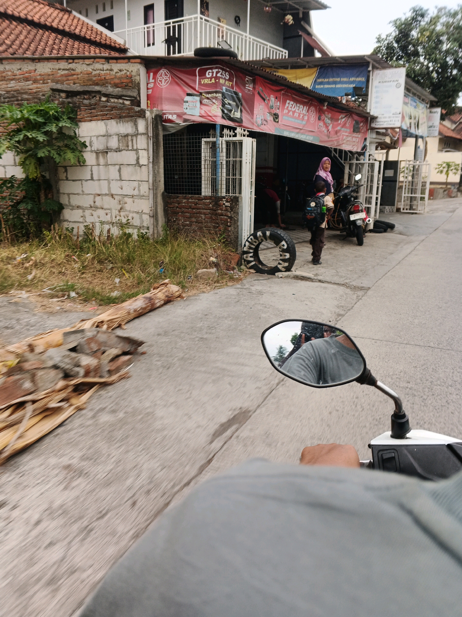 The image shows a street scene in Jangrana, Indonesia. A motorcycle is parked in front of a shop with a red banner. The banner has text in Indonesian and English, including "GT25S VRLA - MF KIT" and "FEDERAL PARTS." A woman wearing a pink hijab and a child with a backpack are standing near the motorcycle. There are tires and other items scattered around the shop. The perspective is from the viewpoint of someone riding a motorcycle, as indicated by the reflection in the side mirror. The environment appears to be a typical street in a town or city, with buildings lining the road and vegetation present. The road surface is paved.