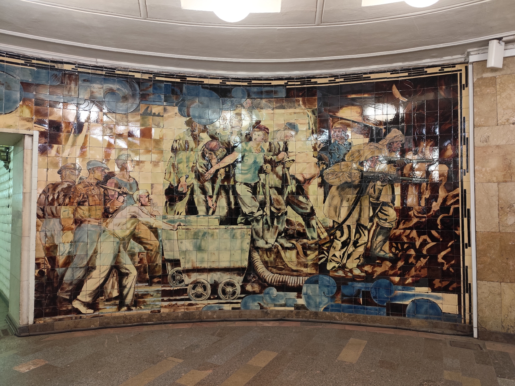 The image shows a large tile mural depicting workers, presumably in a Soviet-era style. The mural is located in what appears to be a public space, possibly a metro station. The style of the artwork is reminiscent of socialist realism.
The mural depicts a group of men in various states of labor. Some are pushing a mine cart, while others seem to be working with tools or machinery. The colors are muted, dominated by browns, blues, and tans, giving the scene a historical feel.
The setting is likely the Moscow Metro, renowned for its elaborate stations featuring artwork and architecture from the Soviet period. The curved shape of the wall, the tiled surface, and the presence of architectural details above the mural are consistent with the architectural style of the Moscow Metro stations.