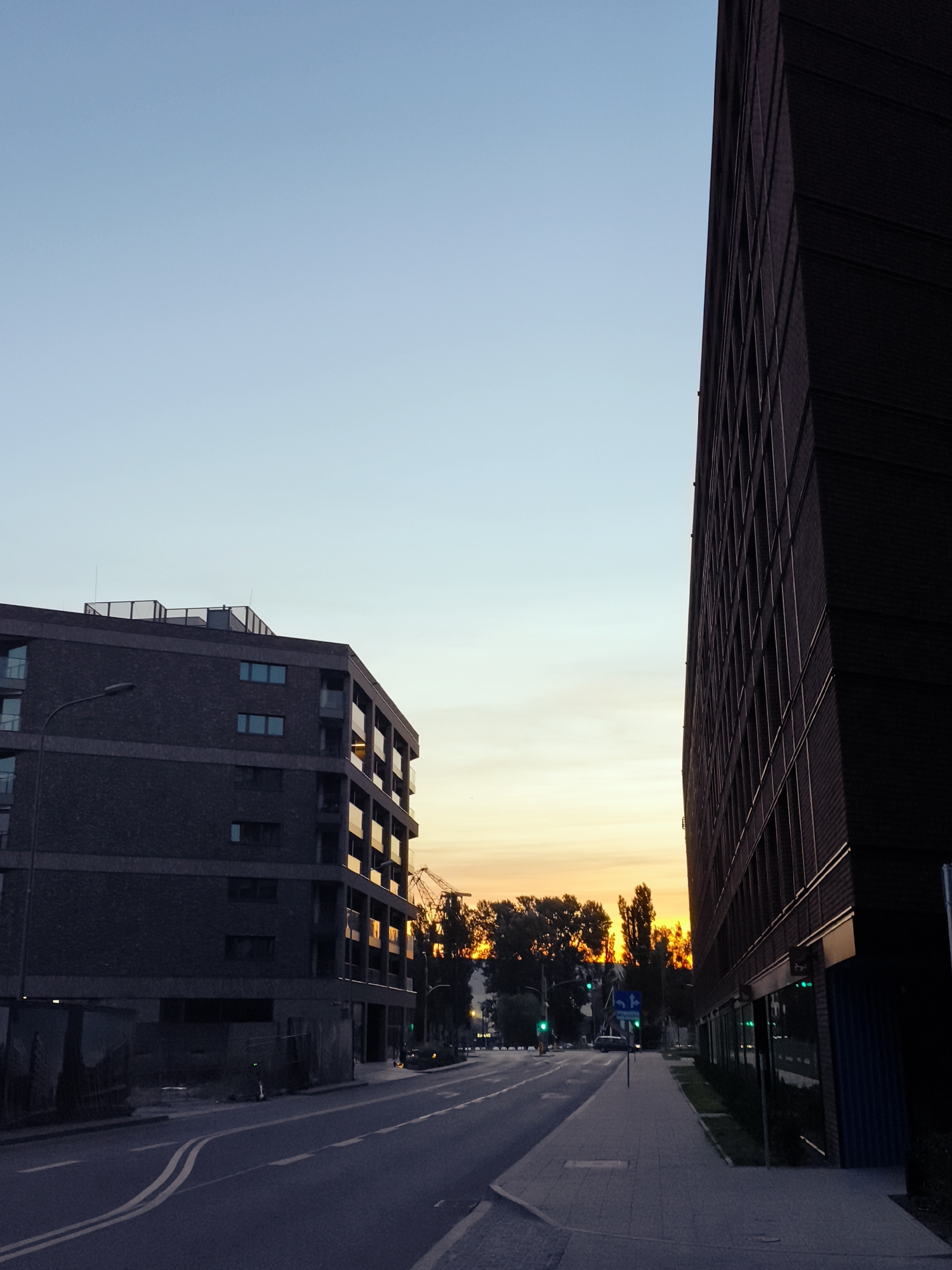 This image captures a street scene in Gdańsk, Poland, at dusk. The sky displays a gradient of pale blue transitioning to a warm orange and yellow sunset near the horizon, highlighting the silhouette of trees and construction cranes. On the left, a modern multi-story apartment building with brick facade and balconies is visible. On the right, a taller, darker building with a brick facade and many windows dominates the foreground. A paved road runs through the center of the image, marked with white lines. A sidewalk runs along the right side of the road. Traffic lights are visible, with some showing green. A few cars and an electric scooter are present on the street and sidewalk. The overall impression is one of an urban environment at the end of the day, with elements of both established architecture and ongoing development.