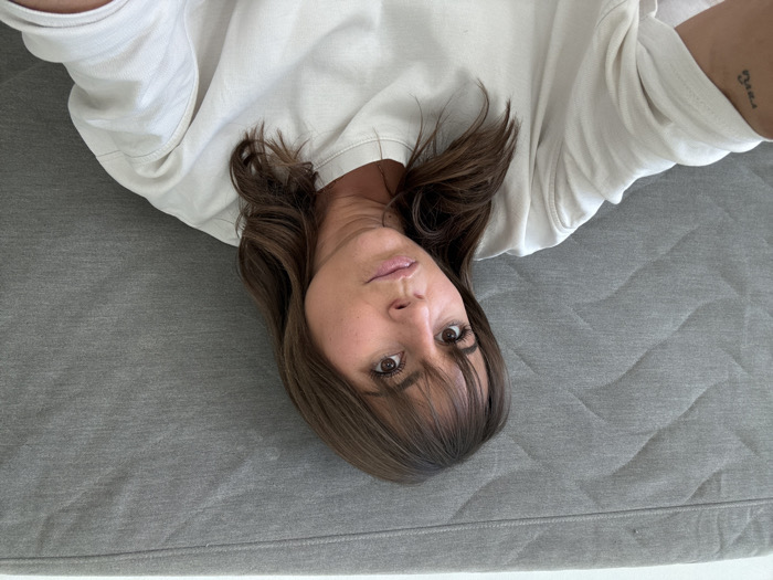 A woman lies on a gray quilted surface, likely a bed or mattress, wearing a white sweatshirt. She has brown hair with bangs and is looking at the camera. A tattoo is visible on her arm. The photo is taken from an overhead perspective, rotated counter-clockwise.