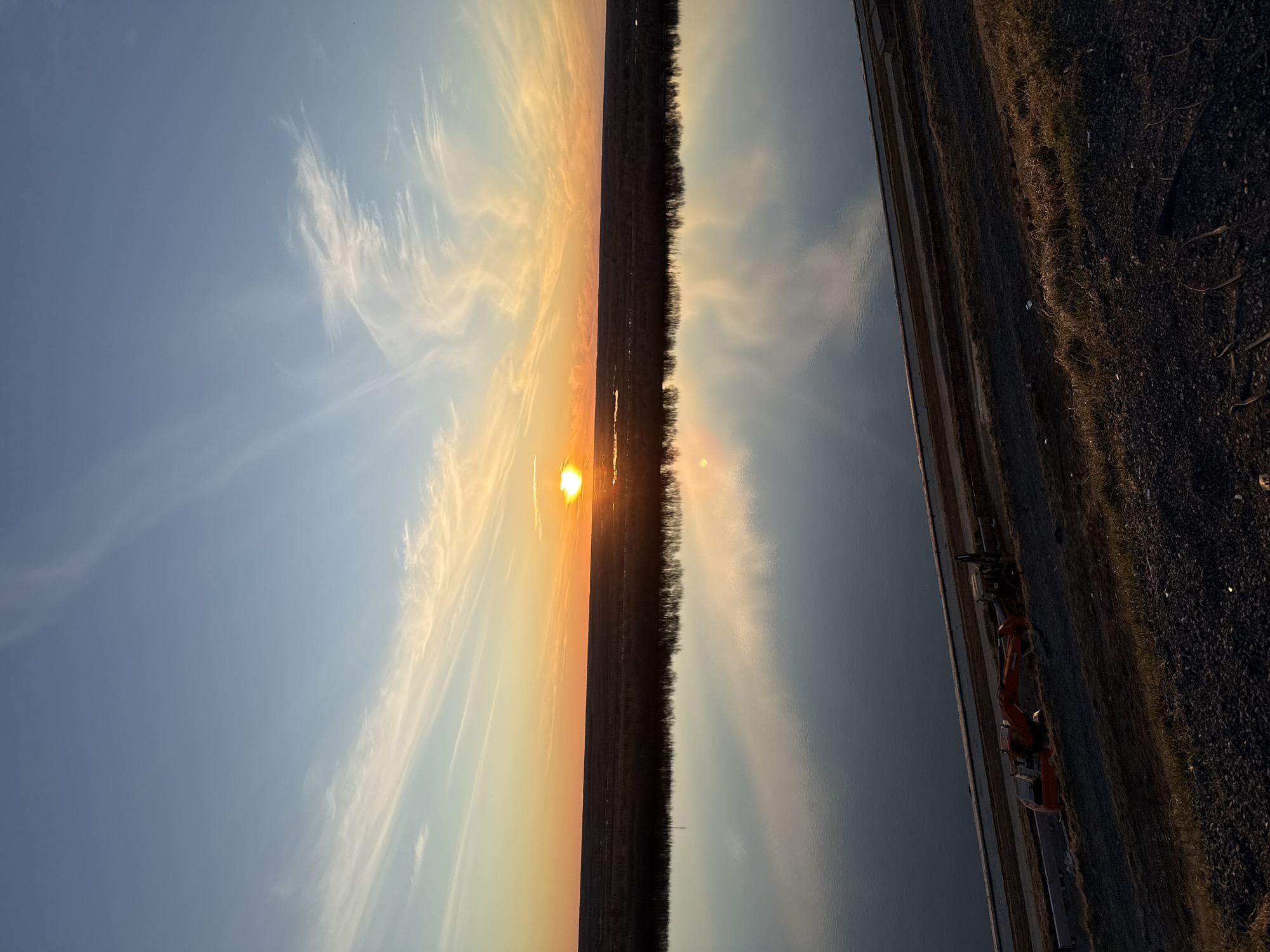 The image captures a serene landscape during either sunrise or sunset, with the horizon line horizontally bisecting the frame. A brilliant, low-lying sun, slightly to the left of center, casts an intense golden-orange light across the sky. The sky transitions from a soft blue in the upper left to vibrant orange and yellow around the sun, adorned with delicate, wispy cirrus clouds that are illuminated by the warm light.
Below the horizon, on the right side, a calm body of water, possibly a river or canal, reflects the subtle light and colors of the sky. A narrow strip of land or a road runs parallel to the water's edge, separating it from a distant, dark silhouette of trees or bushes on the left. The foreground on the right side reveals a gravelly or earthy bank with sparse vegetation. An orange excavator is distinctly visible on this bank, positioned next to the water, with a faint reflection seen in the water's surface.
The scene evokes a sense of tranquility and natural beauty, characteristic of the "golden hour." The presence of the excavator suggests some form of infrastructure work or maintenance near the waterway. There are no visible people or text. The location context suggests this tranquil moment is captured near Tobolsk, Russia.
