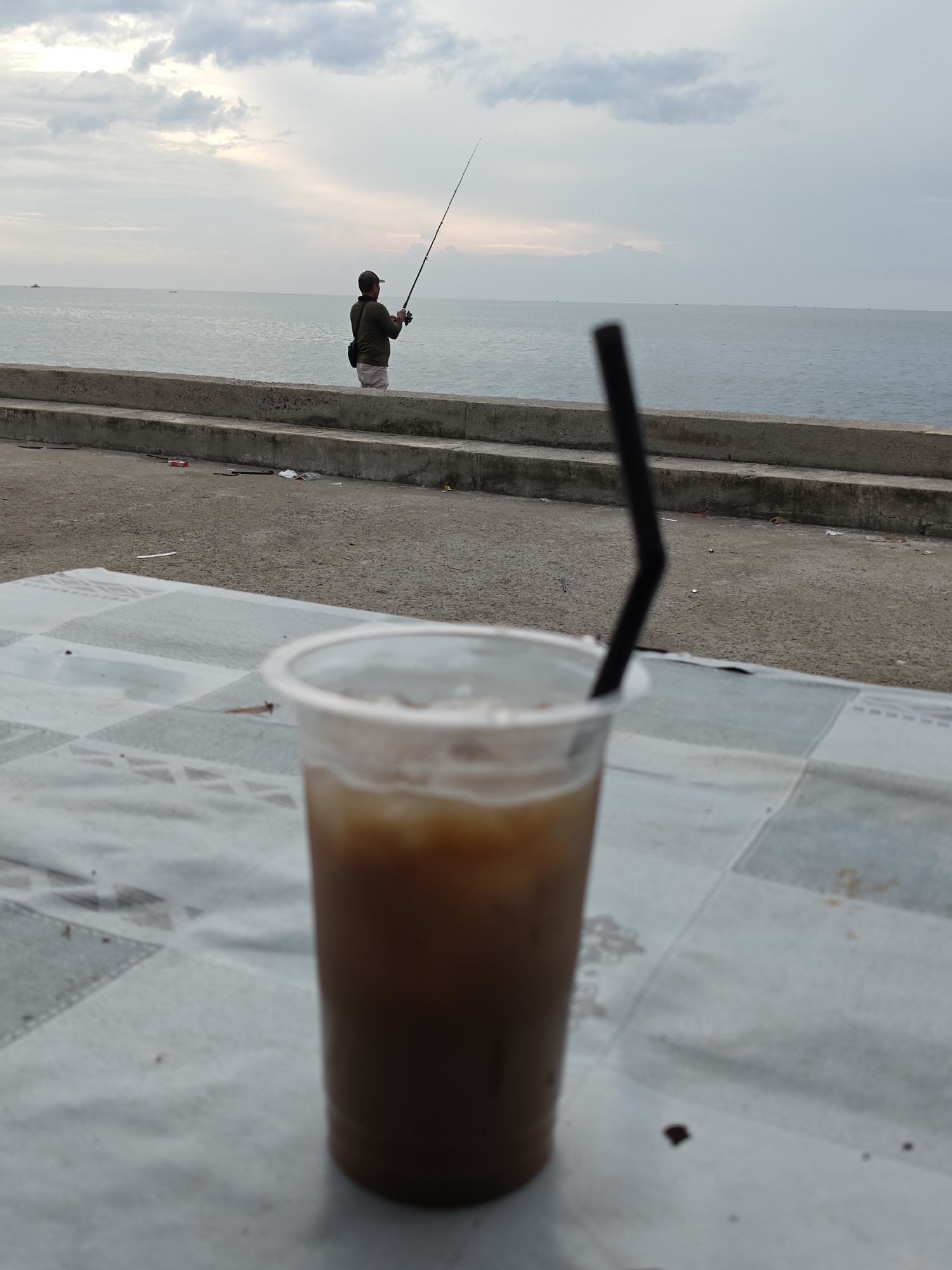 A glass of iced coffee with a black straw sits on a checkered tablecloth in the foreground. In the background, a person fishes by the sea, standing on a concrete barrier. The sky is cloudy, and the sea is calm. The location is the TPI (Tempat Pelelangan Ikan) or Fish Auction Place in Lhokseumawe, Indonesia, a coastal city known for its fishing industry.