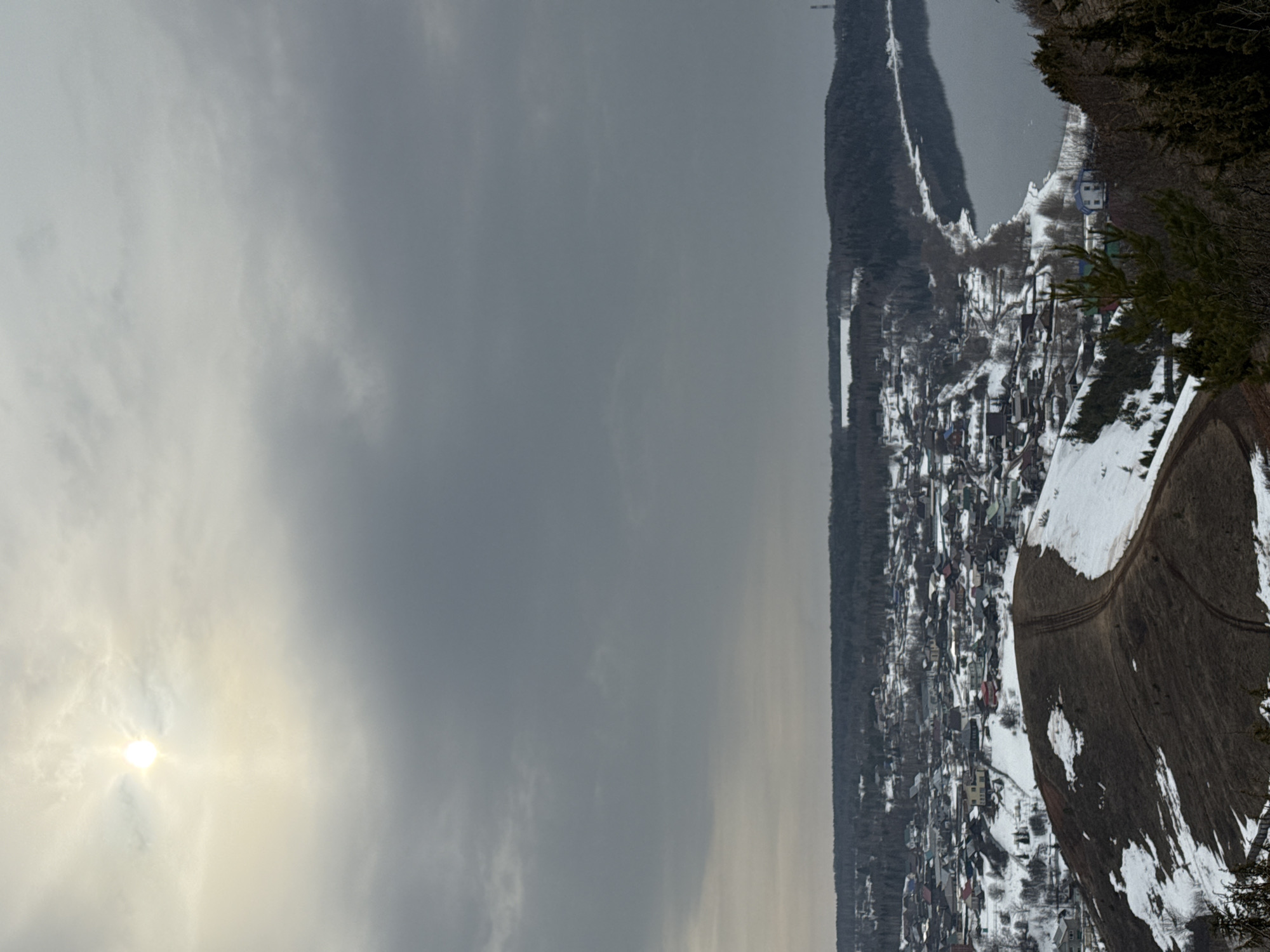 The image presents a panoramic view of the village of Ольховка, Russia, under an overcast sky with a diffused sun visible in the upper left quadrant. The foreground features a steep, barren hillside, primarily dark brown earth with patches of melting snow, marked by two parallel dirt tracks traversing its slope. Sparse, dark green coniferous vegetation is visible at the immediate lower right. In the midground, the village of Ольховка extends across a lower plateau and gentle slopes, characterized by numerous closely clustered residential buildings with varying roof colors (red, blue, grey), many still partially covered in snow. Roads or paths are visible winding through the village. To the right of the village, a wide body of calm water, likely a river, runs along the base of the settlement, reflecting the grey sky. Its shoreline is visible, dotted with bare trees. The background consists of dense, dark forest covering rolling hills, stretching to the horizon. In the far distance, a tall, slender industrial tower and a large, rectangular white building are discernible above the treeline on the right side. No individuals or discernible actions are present in the scene. The environment indicates a late winter or early spring setting due to the partial snow cover and bare deciduous trees.