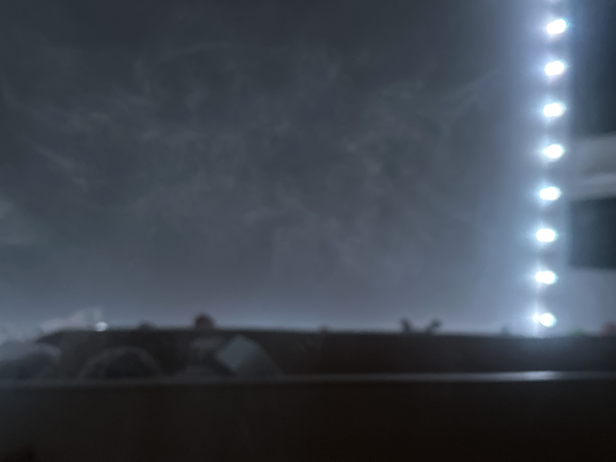 The image displays a dimly lit room, possibly a bedroom, with a large, dark, vertical object on the left side, possibly a wardrobe or a door frame. The right side of the image shows a wall with a hazy, cloudy texture, illuminated by a string of bright white LED lights positioned horizontally along the bottom edge. These lights appear to be decorative, creating a blurred glow. The overall impression is of a domestic interior with minimal detail visible due to low light and a soft focus. There are no discernible people or specific objects that would allow for identification of a location within Petropavlovsk-Kamchatsky, Russia.