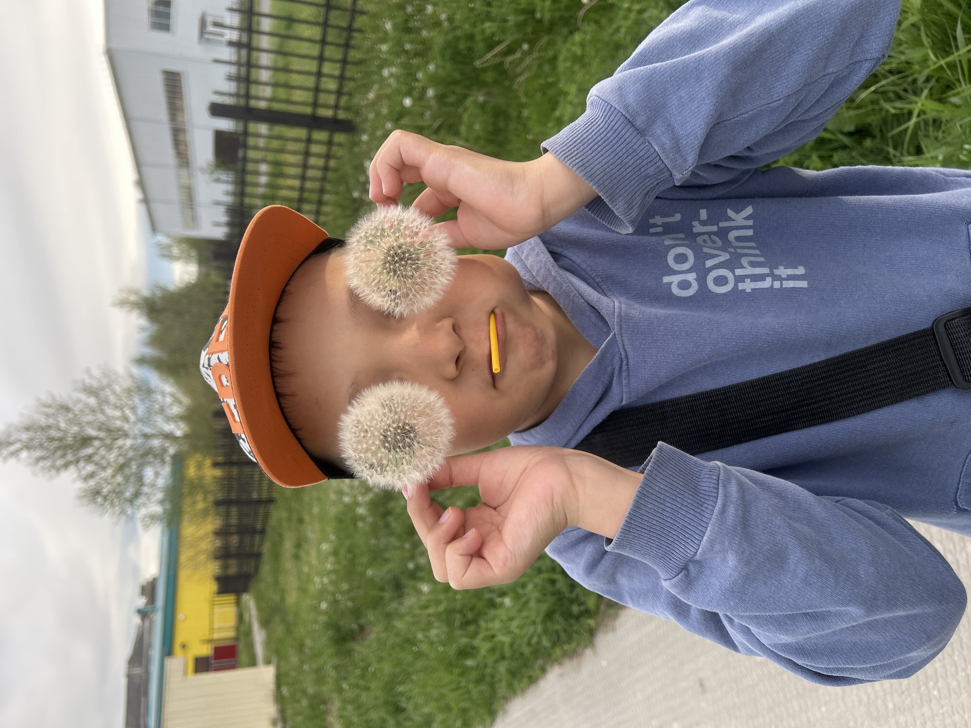 A child is holding two dandelions up to their eyes and smiling with a yellow stick in their mouth. The child is wearing an orange cap and a blue sweatshirt that says "don't over-think it." They also have a black strap going across their body. The background shows grass, a fence, a building, and trees. The image was taken in the city of Uray, Russia.