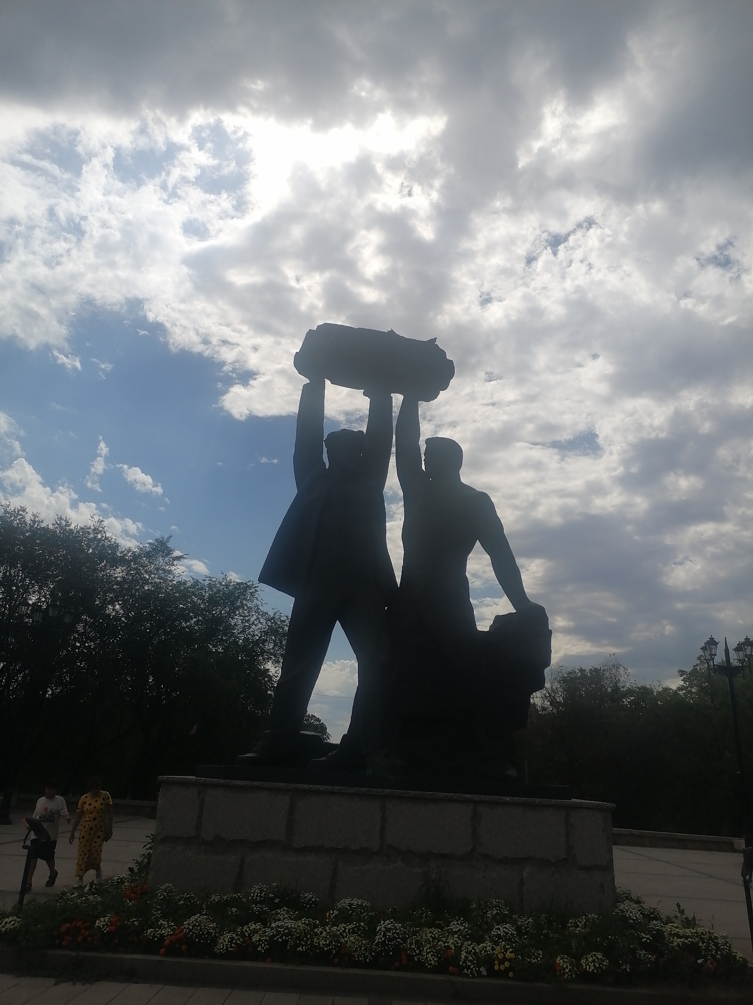 A silhouetted statue of two male figures lifting a large object above their heads stands against a cloudy sky. The statue is on a stone pedestal in what appears to be a park or public square. In the foreground, a flower bed with white and orange flowers is visible. Two individuals, a man and a woman, are walking in the background. The statue is located in front of the city administration building in Qarağandı, Kazakhstan. The sky is partially clear with patches of blue visible between the clouds, and sunlight is breaking through in some areas. Trees and streetlights are also visible in the background.