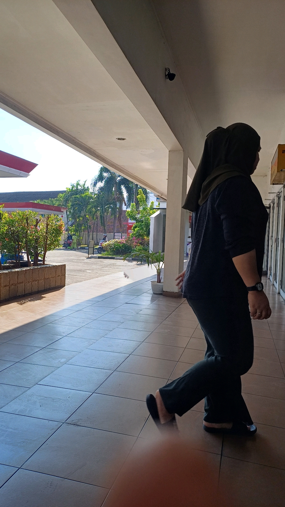 A full-body shot shows a woman wearing a dark green hijab and a dark gray long-sleeved shirt and pants, walking from left to right on a tiled outdoor walkway. She is looking to her right and her left leg is extended forward, her right leg is bent at the knee. Her right hand is visible, resting near her hip. She wears a watch on her left wrist. The walkway is covered by a white overhang. The ground in front of the walkway is tiled with large square tiles. To the left of the walkway, there is a raised planter box with green foliage. Beyond the planter box, palm trees and other greenery are visible, along with a building with a red roof and a gas station sign. The scene appears to be set in an outdoor commercial area. In the upper left, a security camera is mounted on the ceiling.