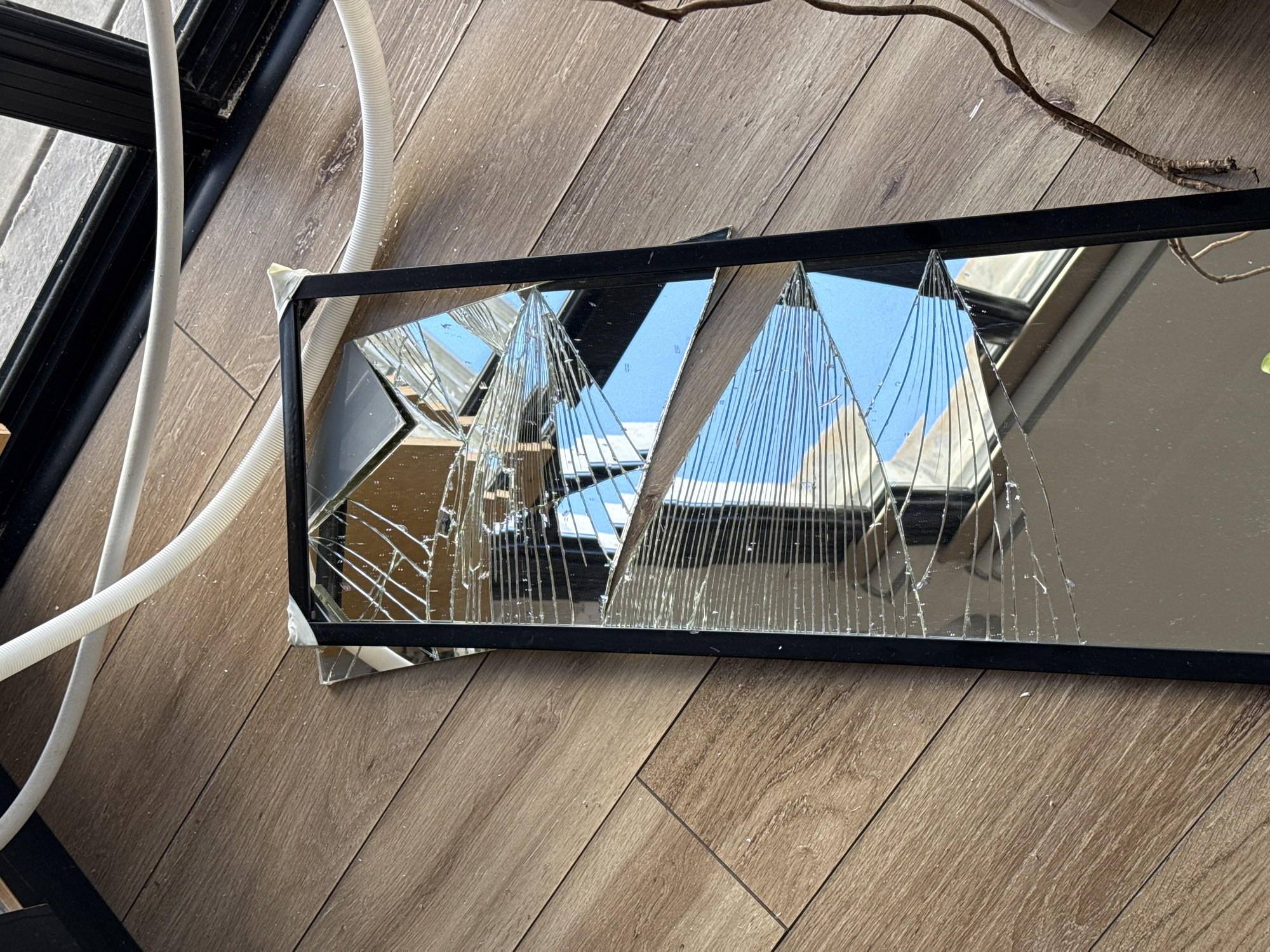 A rectangular mirror with a black frame lies on a light brown wooden floor. The mirror is broken into multiple triangular shards, reflecting an outdoor scene with a blue sky and building structures. To the left of the mirror is a white corrugated tube and part of a black frame. A small branch is visible near the top right corner of the frame. The scene appears to be indoors, possibly a residence. The location is unknown.