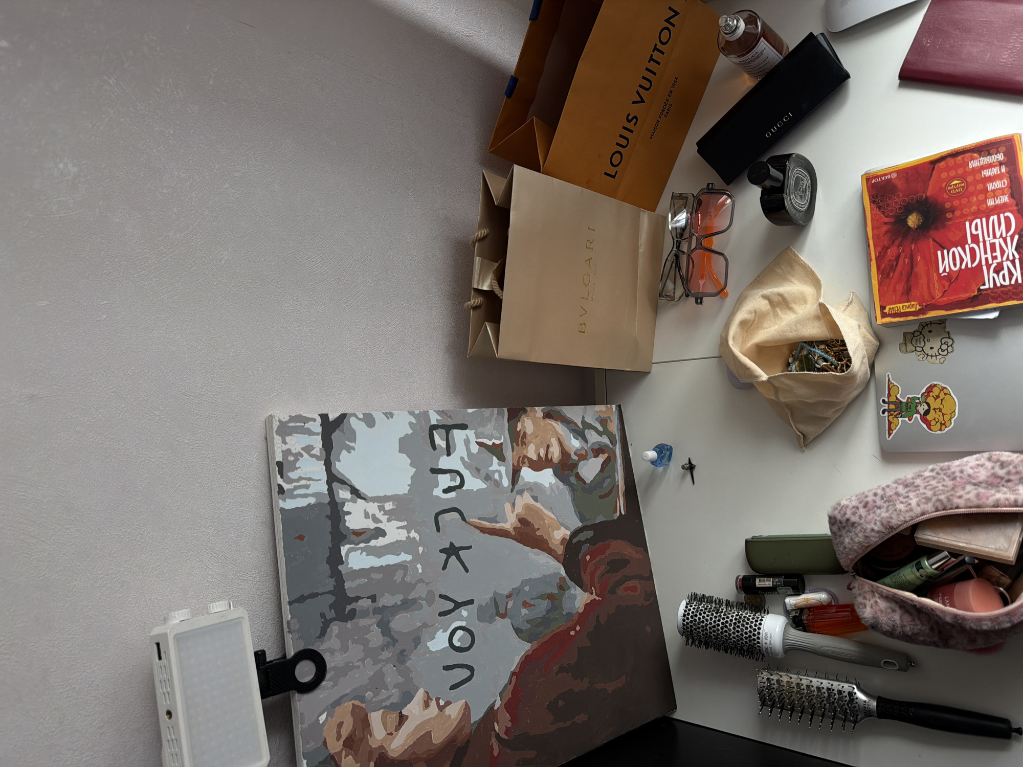 The image depicts a still life arrangement on a white surface, likely a desk or table, against a grey textured wall. The scene is captured from a high-angle perspective, with the lighting source appearing to be from the top left, casting shadows. Dominating the upper left portion of the image is a large canvas with a painting on it. The painting features two figures and appears to have the word "FUCK YOU" written in dark letters across the center. Attached to the top edge of the canvas is a small, white LED light panel. To the right of the canvas, two shopping bags are visible. The left bag is beige with "BVLGARI" printed in gold lettering. The right bag is orange and has "LOUIS VUITTON" printed in black lettering, along with "MAISON FONDÉE EN 1854 PARIS" below it. In the foreground, several objects are scattered on the white surface. There are two hairbrushes, one larger and one smaller, both with black handles. Next to them is a small, orange lighter and a tube of lipstick. A green rectangular object, possibly a small case or battery pack, is also present. A small, pink leopard-print makeup bag is open, revealing various cosmetic items inside. Further to the right, a pair of eyeglasses with clear and orange frames rests on top of another pair of dark-rimmed glasses. A small, light blue bottle, possibly nail polish or a similar liquid, is situated near the painting. A small silver object, possibly an earring or a clasp, is also on the surface. In the mid-right area, a black clutch or case with "GUCCI" printed on it is visible. Next to it is a black perfume bottle with a dark stopper. Another perfume bottle, this one with a glass body and a silver cap, is placed near the right edge of the frame. Towards the bottom right, a book with a vibrant red poppy on its cover is open. Text in Russian is visible on the cover and inside the book. A portion of a red passport or similar booklet is also visible at the far right edge. A small, light-colored pouch, possibly a jewelry bag, is open, revealing a collection of necklaces and other jewelry items. Two stickers are visible on a laptop or similar flat object positioned horizontally in the lower portion of the frame; one depicts a character in a yellow hat, and the