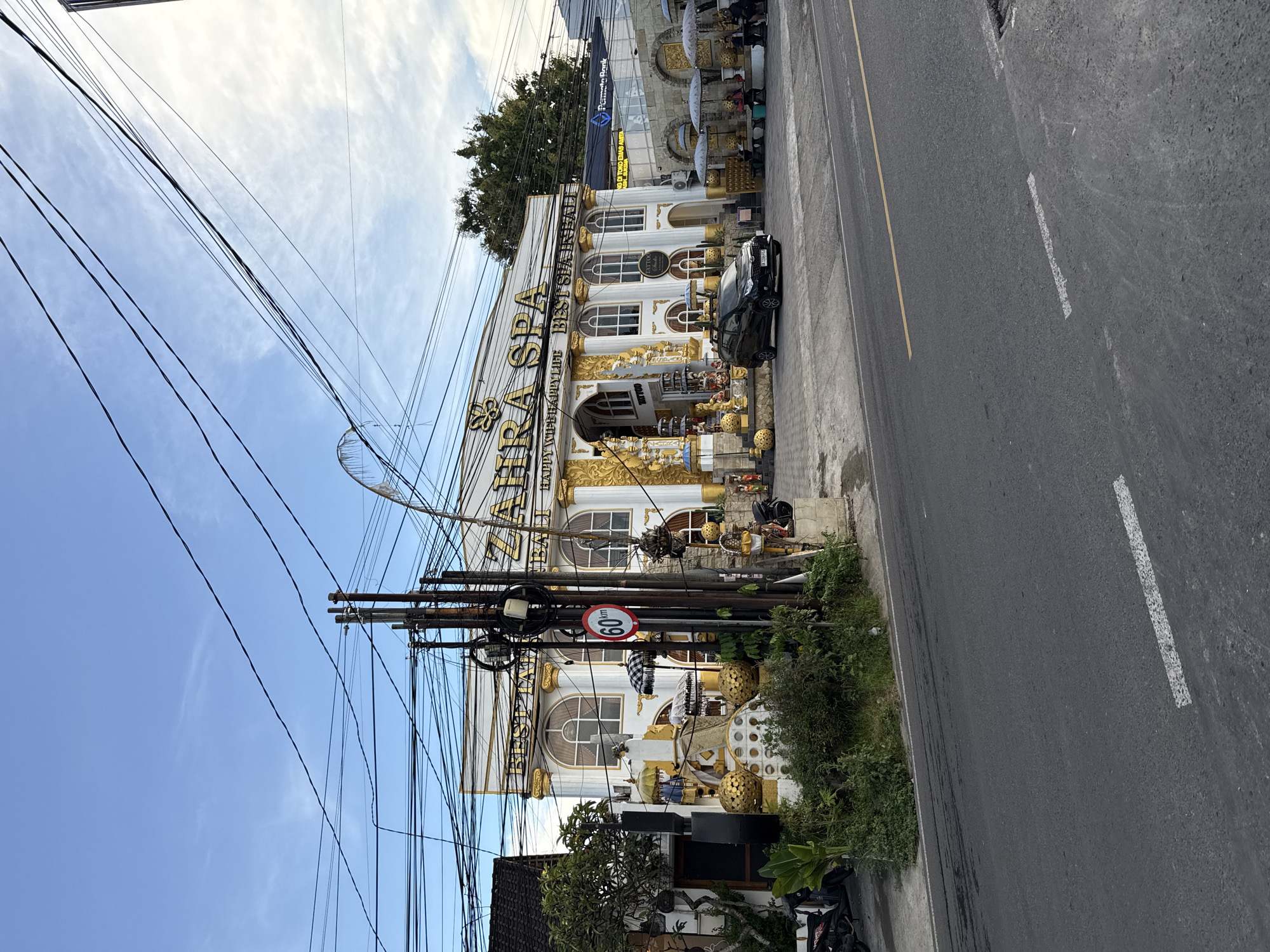 A street-level photograph shows the ZAHRA SPA building in Ungasan, Indonesia, with a black car parked in front and roadside street features.
The building is decorated with gold and white trim with a sign that reads "ZAHRA SPA." There are additional signs indicating "HAPPY WIPE BAPPY LIFE BEST SUAINBAL," along with other unreadable texts.
A black car is parked near the front entrance of the spa. A pole is visible to the side of the spa with numerous electrical cables around it.
The road surface is seen along with a white lane marker to the right of the frame.
The sky is blue with some light clouds.