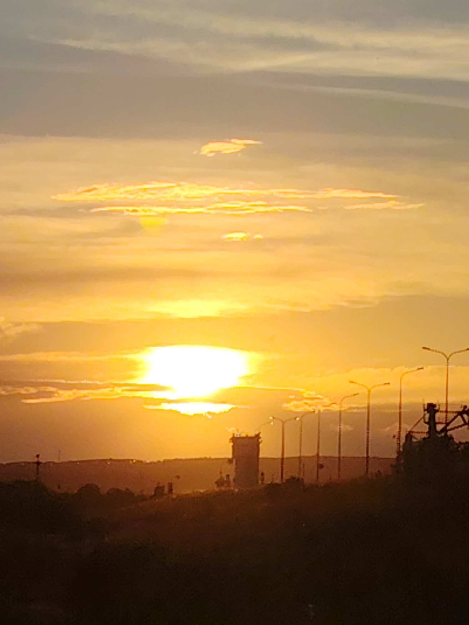 This media file depicts a picturesque sunset over an industrial or port area in Chaikovsky, Russia. The sky is a vibrant spectacle of oranges and yellows, with soft clouds catching the last rays of the sun. The sun itself is a bright, almost blinding orb low on the horizon, casting long shadows and illuminating the scene with a warm, golden light.
In the foreground, the landscape is silhouetted against the bright sky, appearing as dark, earthy tones. Distinguishable elements within this silhouette include patches of vegetation and possibly rolling hills. Further back, a stark, rectangular tower, likely part of an industrial complex or control center, stands prominently. To its right, a series of tall streetlights or lampposts line the horizon, their shapes clearly defined against the illuminated sky. A hint of industrial machinery, possibly a crane or part of a larger structure, is visible on the far right.
The overall mood is one of tranquil beauty, with the natural splendor of the sunset contrasting with the man-made structures. The time of day is clearly evening, as indicated by the setting sun. The weather appears clear with scattered clouds, contributing to the dramatic visual effect of the sunset. There is no visible text in the image. The scene evokes a sense of calm and contemplation, as the day draws to a close.