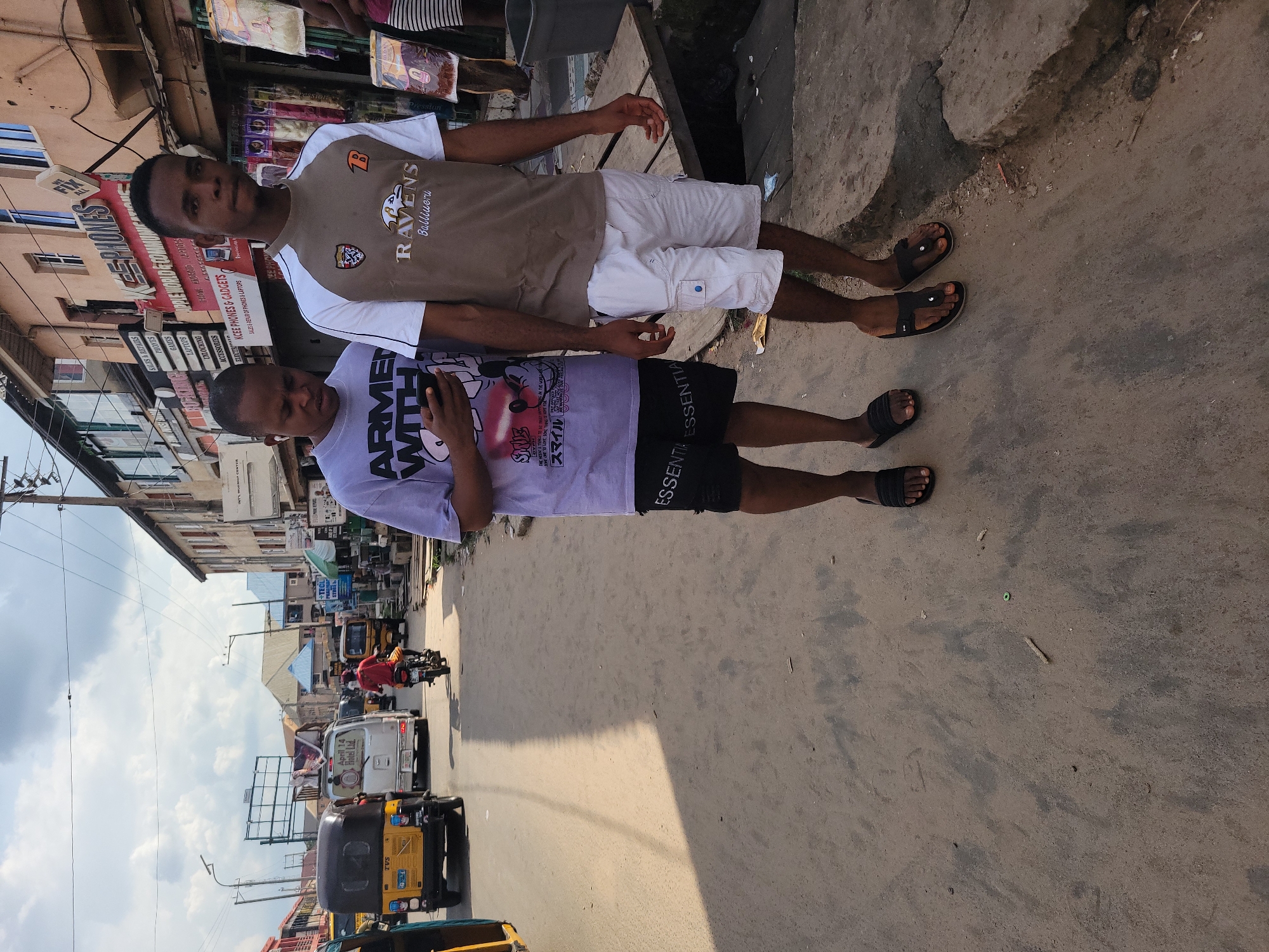 Two young men stand on a dusty, unpaved street in Egbagu, Nigeria. The man on the left, wearing a purple t-shirt and black shorts, holds a smartphone in his hand. The man on the right wears a brown and white t-shirt with "Ravens" and "Baltimore" logos and white cargo shorts. They are positioned in front of shops lining the street. In the background, vehicles, including a yellow auto-rickshaw and a white van, are visible, suggesting a busy urban environment. The sky above is partly cloudy. The scene appears to be a typical street view in a Nigerian city, with both pedestrian and vehicular traffic.