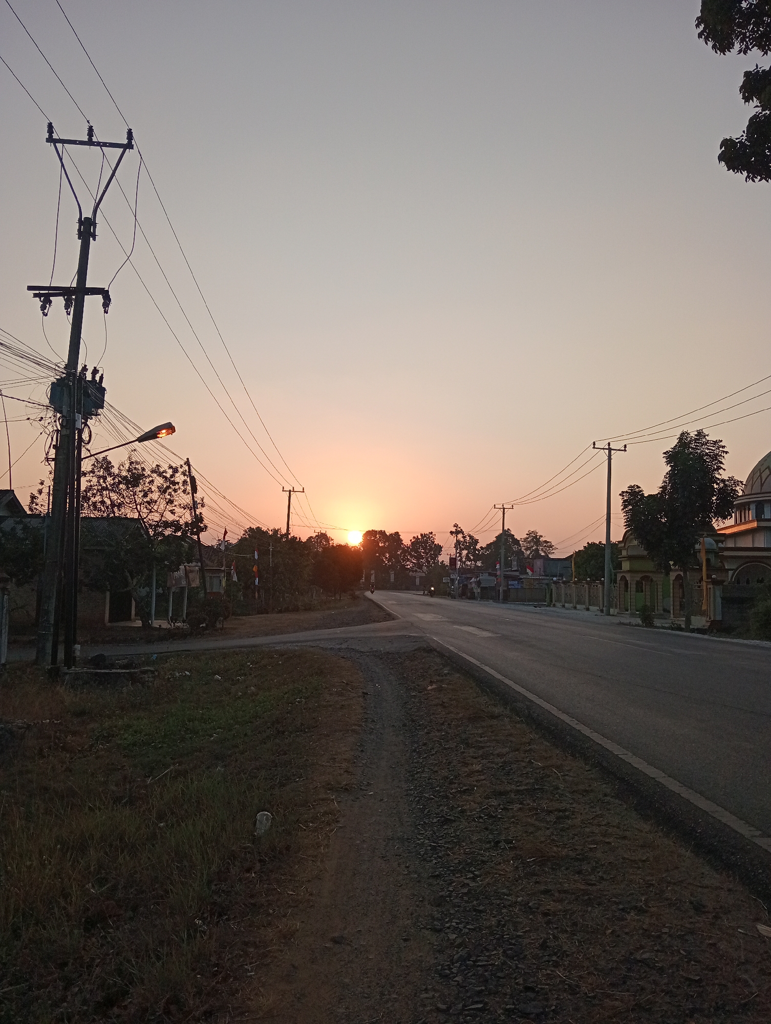 The image captures a serene sunset scene along a road in Taman Negeri, Indonesia. The sky is painted with hues of orange and pink, gradually fading into a pale yellow at the horizon. A paved road, marked with a white line, stretches into the distance, curving slightly to the right. On the left side of the road, a prominent electricity pole with numerous wires and a transformer stands tall. A street light attached to the pole is illuminated. Beyond the pole, several buildings and trees are visible, suggesting a semi-urban or rural residential area. A dirt path runs parallel to the road on the left. On the right side of the road, a mosque with a dome is partially visible, along with a low wall and trees lining the property. The overall atmosphere is calm and tranquil, indicative of the end of the day in a Indonesian setting.