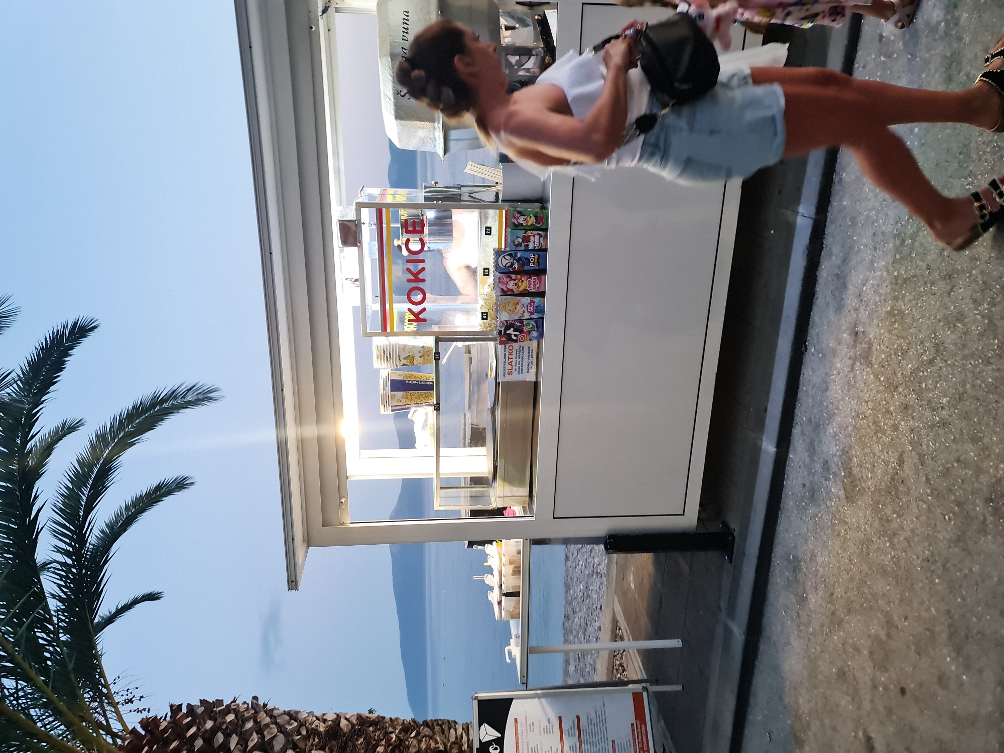 In Općina Gradac, Croatia, a woman in denim shorts and a white top walks past a food stand with the sign "KOKICE" prominently displayed. The stand appears to be selling popcorn and possibly ice cream, with various colorful popcorn bags featuring cartoon characters lined up on the counter. Behind the stand, a body of water is visible, with hills in the distance, suggesting a coastal location. A palm tree stands to the left of the frame, adding to the tropical atmosphere. The scene is illuminated by the artificial lights of the food stand, indicating it is either dusk or night.