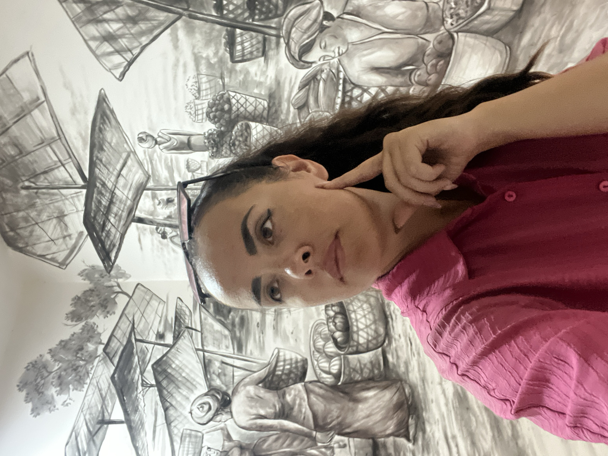 A selfie of a woman with brown hair and brown eyes, wearing a pink collared shirt, is taken in front of a black and white mural depicting a marketplace scene. The mural shows vendors with umbrellas over stalls filled with baskets of produce. The woman is looking off to the side with her left hand touching her cheek. The mural's style suggests a traditional Indonesian setting, consistent with the culture found in locations such as Kuta Selatan, Indonesia, which is known for its vibrant local markets and cultural art.