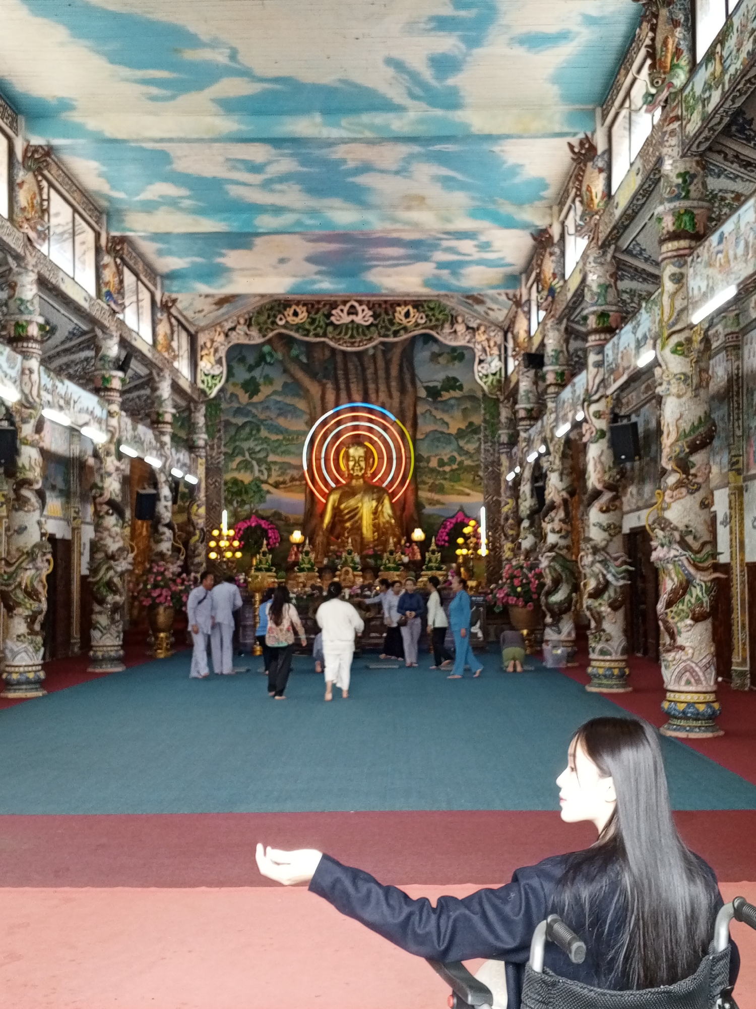 The image depicts the interior of a Buddhist temple in Xuân Thành, Vietnam, characterized by vibrant and ornate decor. A large golden Buddha statue is centrally positioned at the far end of the hall, illuminated by a multi-colored neon halo. The hall's ceiling is painted with a sky and cloud motif. Flanking the central area are elaborately decorated pillars, featuring intricate carvings and figures, possibly depicting mythological creatures or deities. The floor is covered with a large carpet, divided into a teal section in the middle and a maroon border.
Several people are visible within the temple. In the foreground, a young woman with long black hair is seated in a wheelchair, her left arm extended and hand open as if in a gesture of offering or supplication. She is looking towards the right of the frame. In the middle ground, a group of individuals are walking. Some are dressed in white, possibly monks or devotees, while others wear casual clothing. They appear to be moving towards or away from the Buddha statue. The overall atmosphere seems serene, with the architectural and artistic elements contributing to a spiritual and visually rich environment.