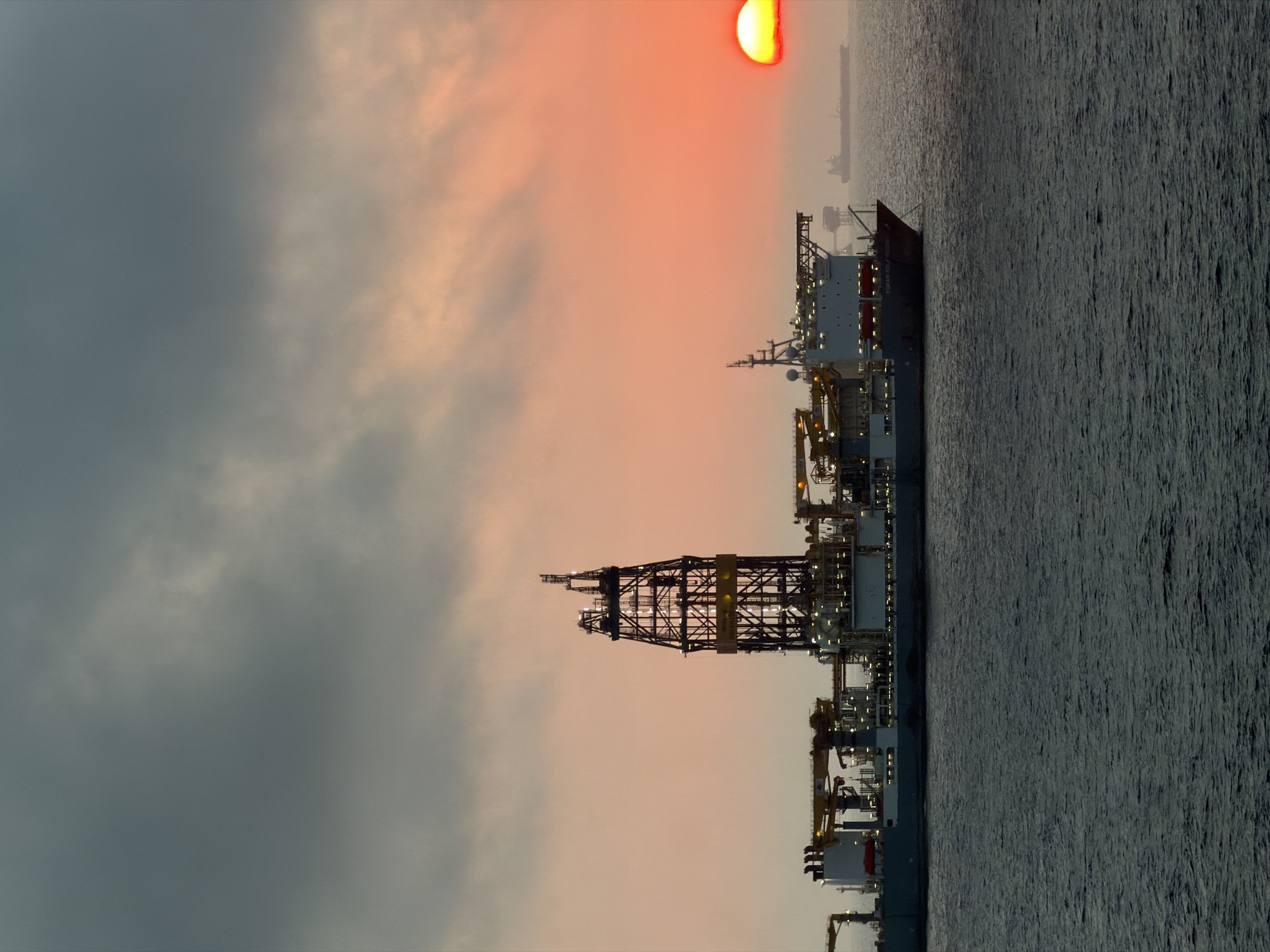 The image captures a dramatic scene of a large industrial ship, likely an offshore drilling rig or specialized vessel, at sea during either sunrise or sunset. The sun, a bright and intense orange-red, glows intensely low on the horizon in the upper right, partially obscured by soft clouds.
The sky exhibits a gradient of colors, transitioning from a darker, cloudy grey on the left to warm, soft pink and orange hues around the sun. The water, which dominates the lower portion of the frame, is a dark, textured expanse of choppy waves, reflecting the low ambient light.
The primary subject is the massive ship, positioned across the mid-ground. It features a prominent, tall derrick structure in the center, characteristic of offshore drilling or heavy lifting. The vessel's decks are elaborate with multiple levels, cranes, and various equipment, many parts of which are illuminated by bright artificial lights. Its dark hull stands out against the warmer sky. A smaller, distant vessel or structure can be faintly seen on the horizon further to the right.
On the stern section of the main ship's hull, white text is visible. The word "ROWAN" is clearly legible, followed by another partially obscured word that appears to be "RENAISSANCE", suggesting the vessel's identity. The scene portrays a powerful blend of industrial might and the natural beauty of a coastal or open ocean twilight.