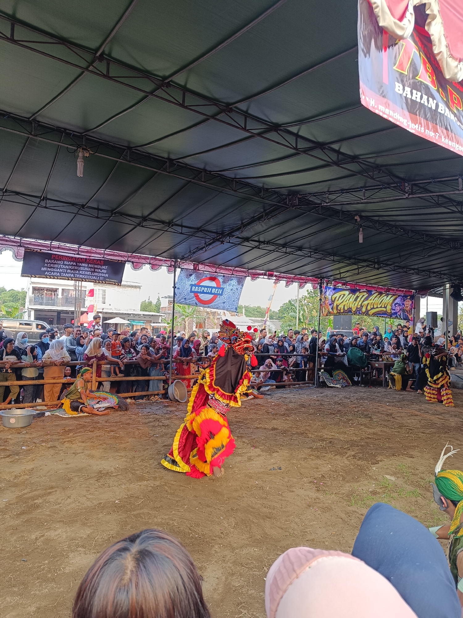 This image depicts an outdoor performance in Jetis, Indonesia, likely a cultural event or festival. The scene is set under a large, green tarp supported by a metal frame, providing shade. In the foreground, a performer in a vibrant, traditional costume with red, yellow, and black accents is captured mid-dance. The costume includes elaborate headwear and flowing garments. To the left and behind the performer, an audience is gathered, seated and standing behind a wooden fence.
Several banners are visible. One, on the left, is black with white text that appears to be a warning or announcement. Another, in the center, displays the text "RASPIJI BEJI" within a red circle and white background. To the right, a colorful banner with the text "TRANT ANOM" is prominently displayed, featuring stylized lettering and imagery.
The ground is covered in sand or dirt, suggesting an informal or rural setting. In the middle ground, several individuals are seated on the ground, some appearing to be participants or organizers. A person wearing a green and yellow headdress is visible on the right side of the frame. In the background, buildings and trees can be seen, indicating an urban or semi-urban environment. The overall impression is one of a lively community gathering centered around a traditional performance.