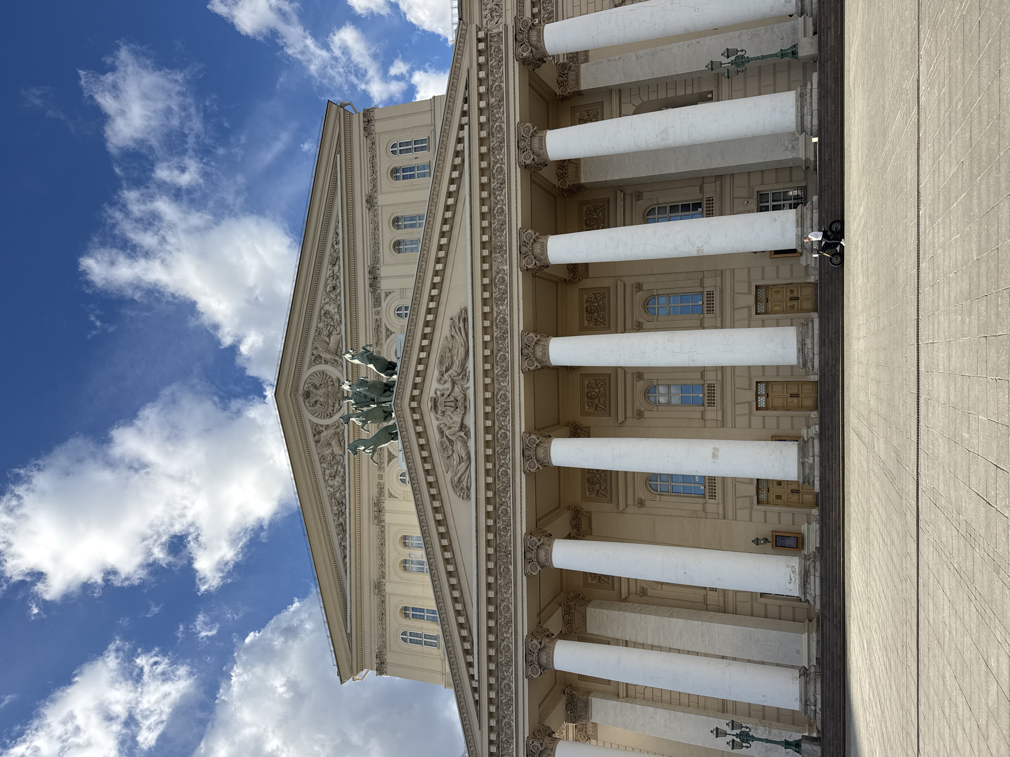 The image shows the Bolshoi Theatre in Moscow, Russia, on a sunny day with a blue sky dotted with white clouds. The grand facade of the theatre, characterized by its neoclassical architecture, is prominently displayed. A series of large, white Ionic columns support a pediment with intricate sculptures, including a chariot pulled by horses at the apex. Below the pediment, a frieze runs along the building, adorned with decorative relief work.
In the foreground, a paved plaza stretches out towards the theatre's entrance. A person on an electric scooter is captured in motion on the plaza, to the right of the central steps leading up to the theatre. Two ornate street lamps stand on either side of the plaza, flanking the entrance area. The overall scene conveys a sense of grandeur and cultural significance associated with the iconic Moscow landmark.