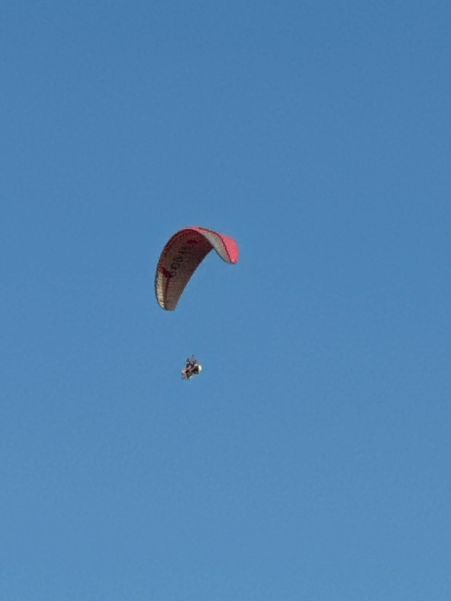 A powered paraglider with a red and grey wing is in flight against a clear blue sky. The paraglider is positioned in the upper left portion of the frame. Below the paraglider, a small pilot harness with a pilot is visible. The overall scene depicts an aerial activity without any discernible urban or natural features that would suggest a specific location within Boreti, Montenegro.